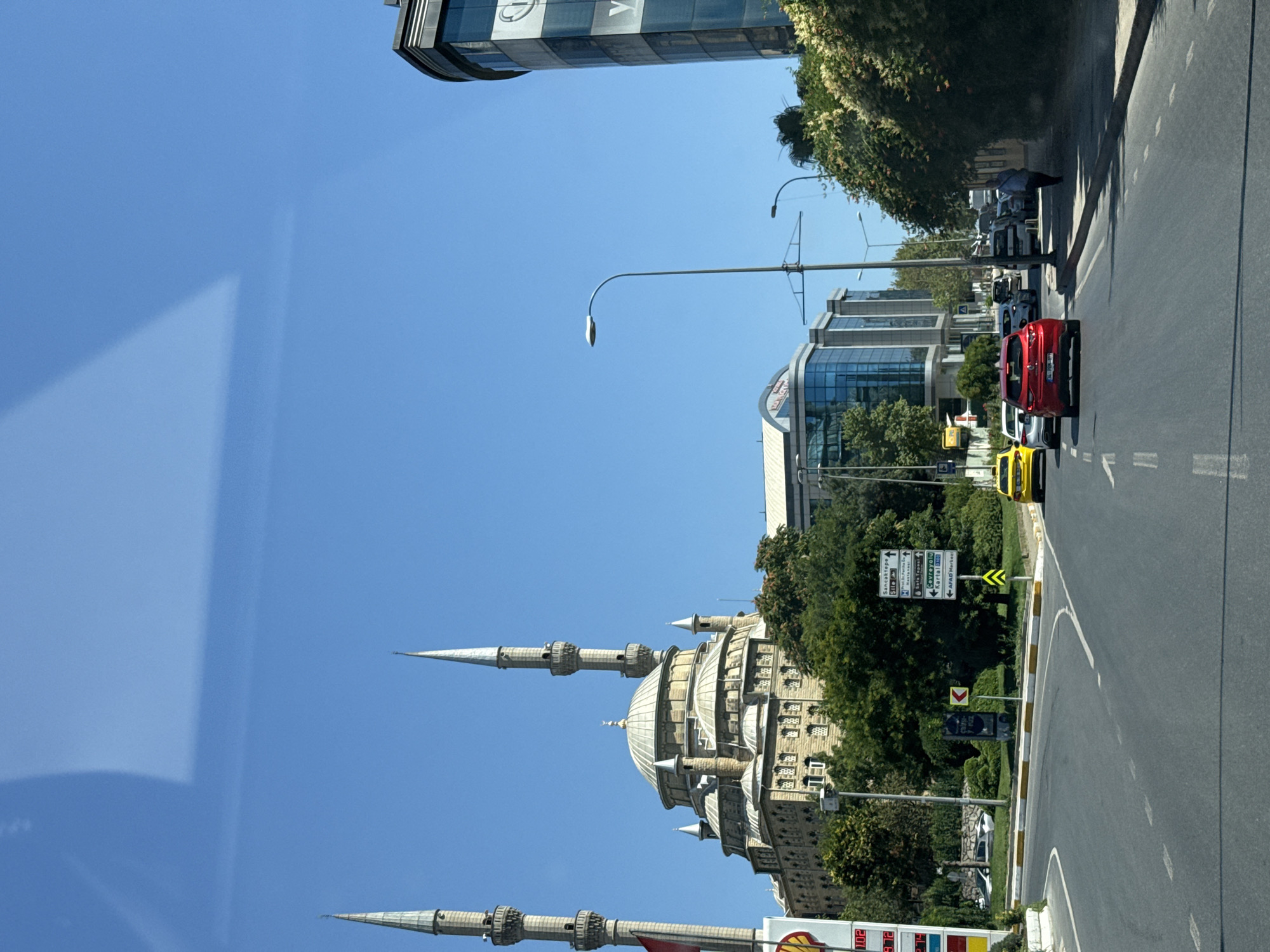 The image depicts a street scene in Sancaktepe, Turkey, under a clear blue sky. In the background, a prominent mosque with multiple domes and minarets stands behind a row of trees. Adjacent to the mosque is a modern glass-fronted building. On the right side of the road, another multi-story building with a curved glass facade is visible. The foreground shows a road with several cars, including a red car and a yellow taxi. Street lights and traffic signs are also present. The scene appears to be a sunny day in an urban environment with a mix of traditional and modern architecture.
