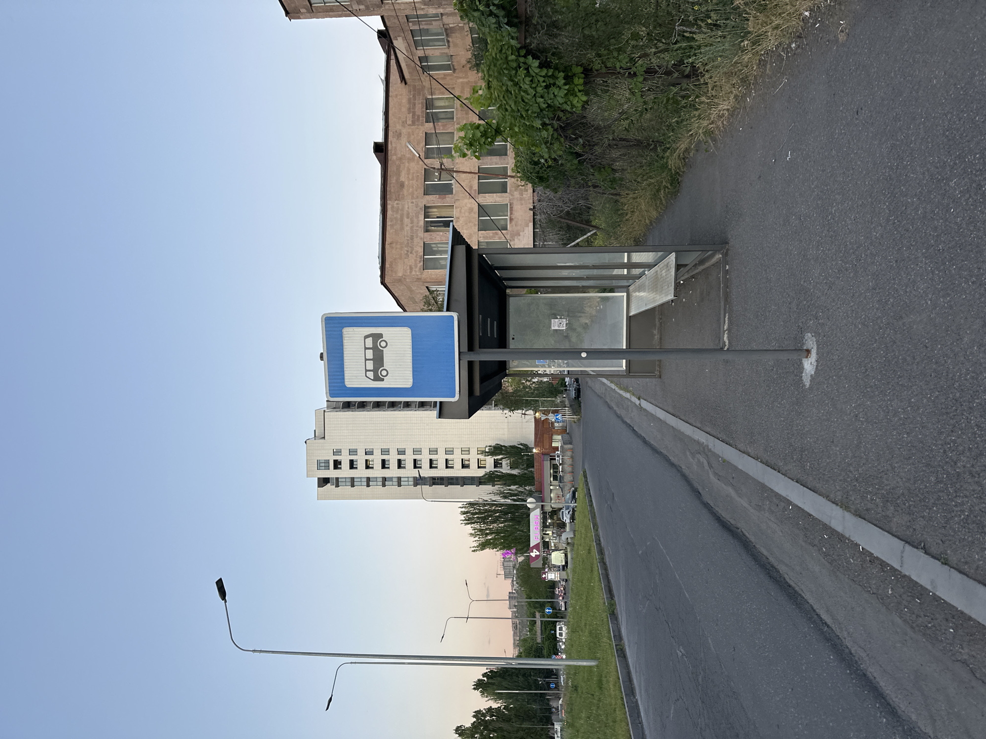 The image depicts a roadside scene in Yerevan, Armenia. A bus stop is prominently featured, marked by a blue sign with a white bus icon. The bus stop has a metal frame with glass panels, one of which has a small poster. Adjacent to the bus stop is a paved road with a curb, showing some signs of wear and possible wetness.
In the background, various buildings are visible, including a tall, modern building with numerous windows and an older building made of reddish-brown stone. Green trees and other vegetation line the road and fill the scene in the distance. Streetlights are positioned along the road, extending into the horizon, suggesting an urban setting. The sky appears clear, indicating fair weather conditions. No people are visible in the immediate vicinity of the bus stop. The scene portrays a typical urban street environment in Yerevan, Armenia.