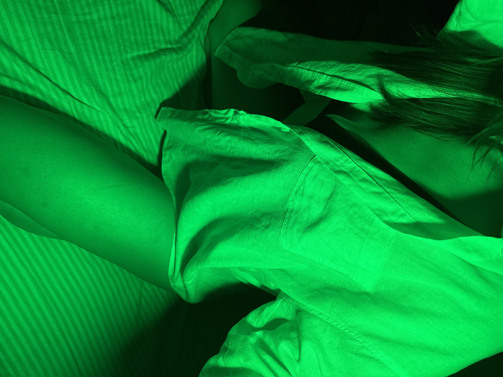 A person lies on a striped bedsheet, dressed in a button-down shirt. Hair is visible in the upper right of the frame. The scene is illuminated with green light. There is no indication the image was taken in the city of Chaikovsky, Russia.