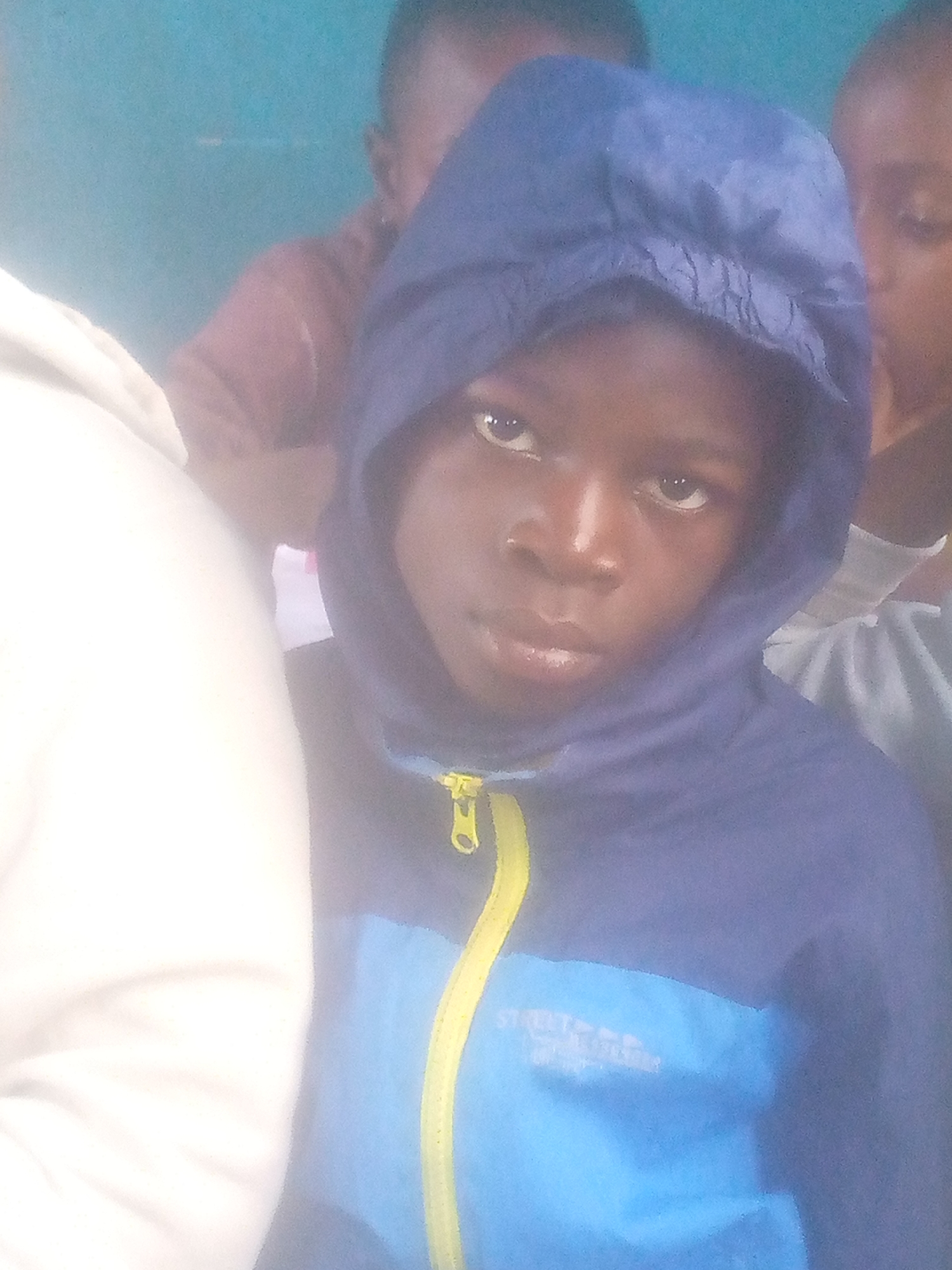 A child is wearing a blue hooded jacket with a yellow zipper. The bottom half of the jacket is a lighter blue. There are other children partially visible in the background. It appears the photo was taken indoors. No further information about the location is discernable from the image.
