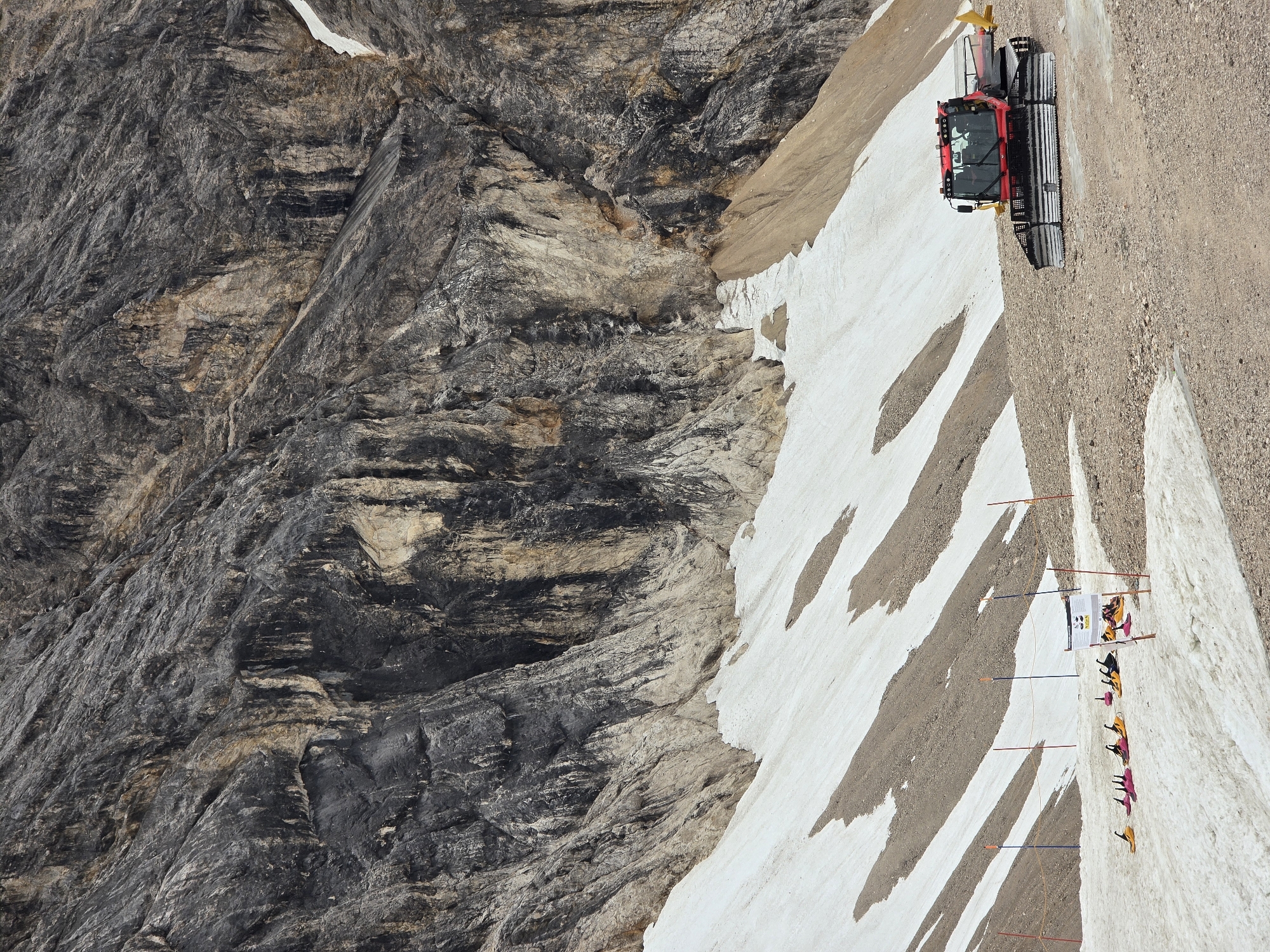 This is an aerial, landscape-oriented photograph taken on a sunny day, likely in the afternoon given the shadows, at a mountainous location in Garmisch-Partenkirchen, Germany. The scene depicts a steep, rocky mountainside with patches of snow. Dominating the right side of the frame is a snow groomer, a red vehicle with a yellow attachment at the rear, positioned on a gravelly slope adjacent to a band of snow.
To the left of the snow groomer, a series of brightly colored flags, primarily yellow, pink, and orange, are planted in the snow, marking a course or area. A few individuals are visible near the flags, some wearing brightly colored jackets, suggesting they might be participants or organizers for an event. One person appears to be standing near a small white structure or board displaying some information, with what looks like a collection of bags or equipment nearby.
The left side of the image is dominated by the rugged texture of the mountain face. Layers of dark grey and lighter brown rock are visible, showcasing the geological formations of the area. The contrast between the barren rock, the scattered snow, and the man-made elements like the snow groomer and flags creates a dynamic and stark landscape. The overall impression is one of an outdoor activity or event taking place in a challenging, high-altitude environment.