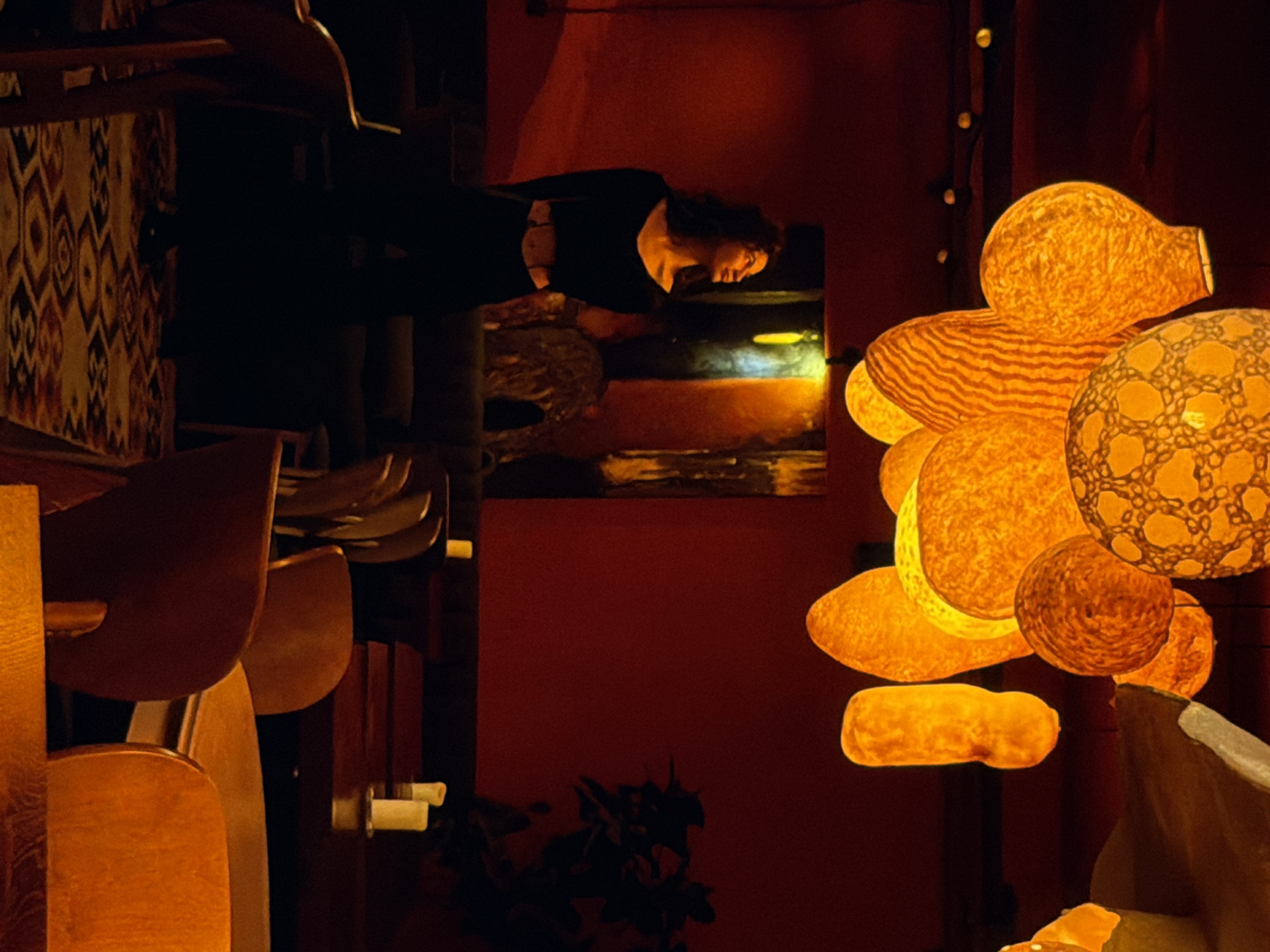 A dimly lit interior in Đà Nẵng, Vietnam is depicted. A person wearing dark clothing leans over a painting hanging on a dark red wall. Nearby, a cluster of irregularly shaped, illuminated orange orbs hangs. In the foreground, a row of empty wooden chairs is visible. A patterned fabric drapes over the back of one chair.