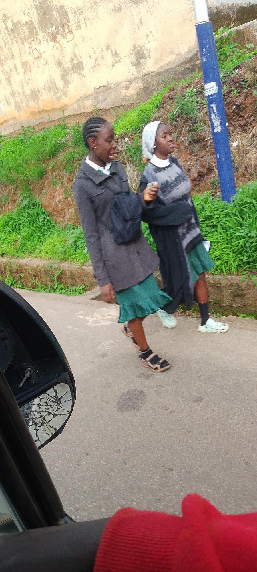 Two young girls, appearing to be students based on their attire, walk along a paved road. The girl on the left wears a dark grey coat over a white shirt and a green skirt, with a black backpack slung over her shoulder. She has braided hair and is looking to her right with her mouth slightly open. The girl on the right wears a grey sweater with a dark pattern over a white top and a green skirt. She has a light-colored headscarf and is looking forward. They are walking on a road with some water stains and debris. Behind them, a steep embankment is covered in green grass and vegetation. A blue pole stands on the right side of the road, and a textured, light-colored wall with red graffiti reading "BLOCK B" is visible in the background on the left. A cracked side mirror of a vehicle is in the lower left corner of the frame, and a red fabric object, possibly clothing, is visible in the lower foreground. The scene appears to be an outdoor setting, possibly in an urban or semi-urban area. Given the context of the graffiti and the general appearance, the location is consistent with the city of Jos, Nigeria, known for its hilly terrain and student population.