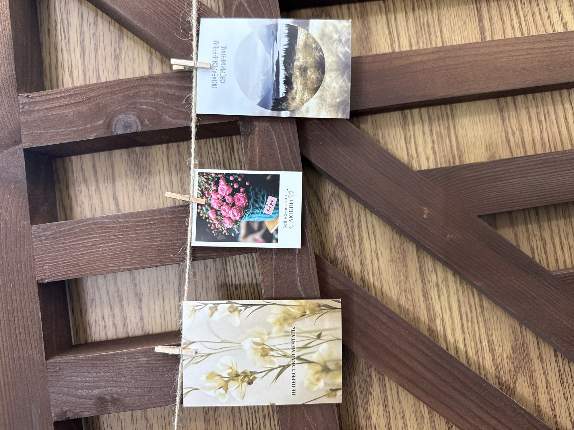 Three printed cards are hanging from a twine string, suspended by wooden clothespins against a wooden lattice structure. The card on the left features an illustration of pale yellow flowers with the text "НЕ ПЕРЕСТАВАЙ МЕЧТАТЬ" (Don't stop dreaming) in Russian. The middle card displays a photograph of a turquoise basket filled with pink roses, with a "For you" tag, and the Russian text "Всё начинается с любви" (Everything begins with love). The rightmost card shows a scenic photograph of a lake and mountains, with the Russian text "ОСТАВАЙСЯ ВЕРНЫМ СВОИМ МЕЧТАМ" (Stay true to your dreams). The background is a light-colored wooden wall with visible wood grain. The overall scene suggests a decorative display, potentially in a home or studio setting. There are no visible people or animals. The image was captured in the city of Votkinsk, Russia.