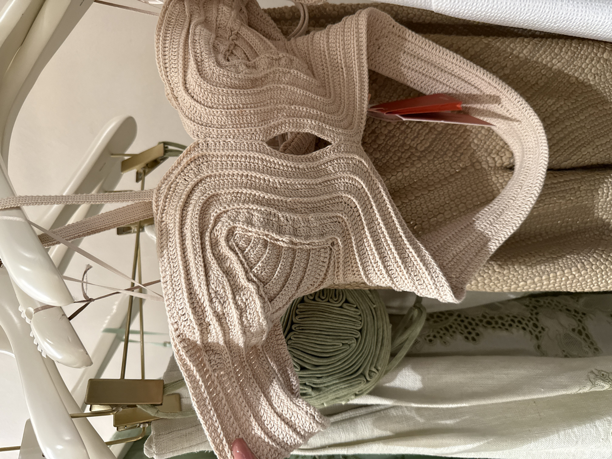 The image shows a beige knitted top on a hanger, hanging among other items in what appears to be a retail setting. The top has a unique, almost architectural design, with concentric rows of knitting creating a layered effect. A price tag is partially visible on the top. Next to the top is a roll of green fabric. Beneath those items are layers of other fabrics, including a cream-colored textured material, and white fabric. The image was taken at an indoor location in the city of Sunrise, United States of America.