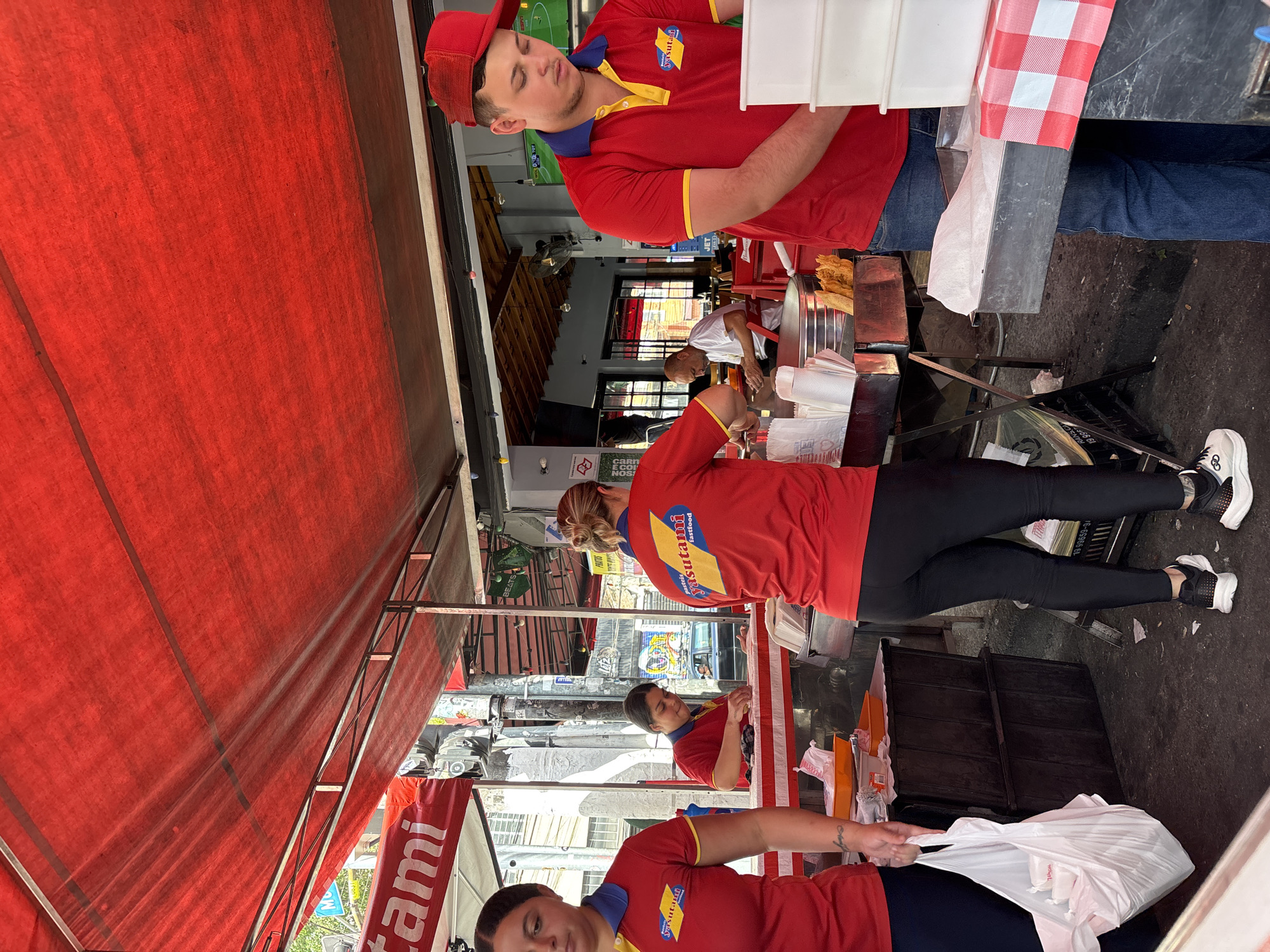 This street scene shows at least three people wearing red and yellow uniforms, possibly for a food stand named Yasutami Fastfood. They are under a red awning, seemingly preparing food or packaging orders. The man in the foreground is wearing a red cap and jeans. The woman in the center is wearing black leggings and sneakers. A plastic bag is being held open, presumably to place items inside. In the background, people can be seen inside the building. Based on the banner visible, this is in São Paulo, Brazil.
