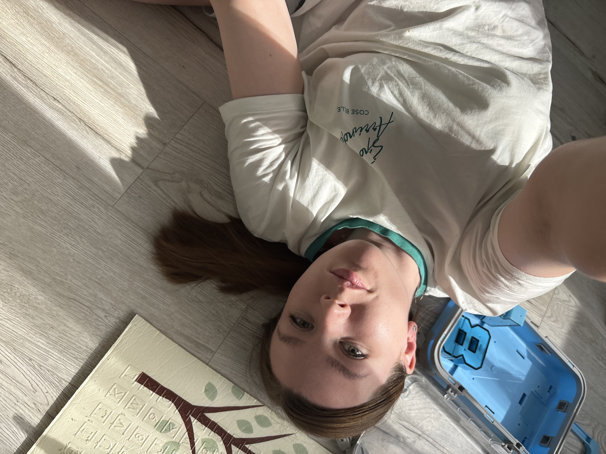A woman lies on her back on a light-colored wood-patterned floor. She is wearing a white t-shirt with green trim around the neck. Her brown hair is spread out on the floor. To her left is a play mat with a tree design and letters/symbols. To her right is a light blue plastic container. The environment is lit by sunlight casting shadows on the floor. The location in the city of Izhevsk, Russia, is not evident.
