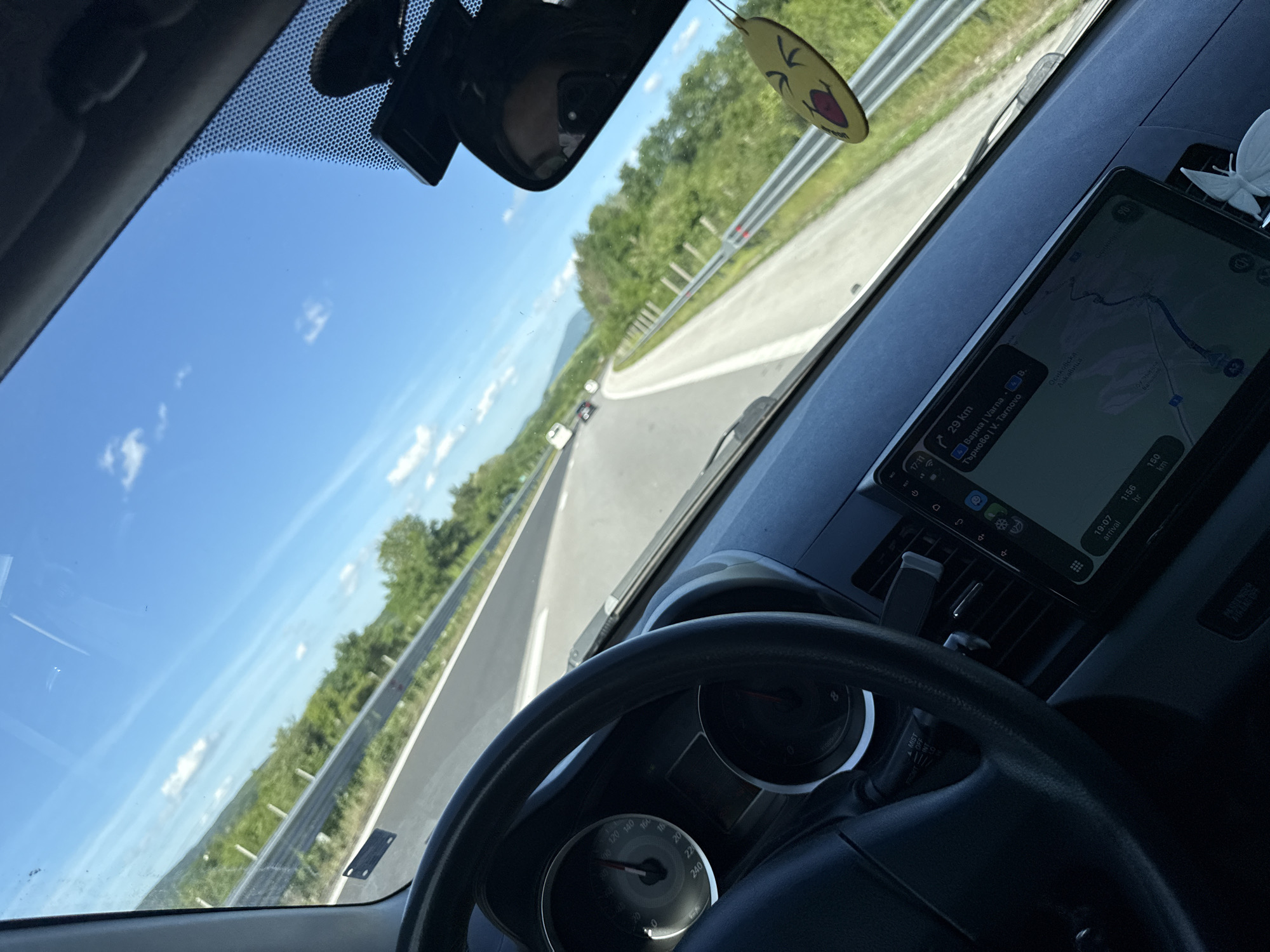 The image is taken from inside a car, looking out through the windshield onto a highway. The sky is blue with scattered clouds, and the road is flanked by trees. A navigational device shows a route and indicates that the destination is 29 km away, displaying 'Varna' (a Bulgarian city) and 'V. Tarnovo' (Veliko Tarnovo, another Bulgarian city). The image is captured while in the location of Osikovska Lakavitsa, Bulgaria. A steering wheel and part of the car's dashboard are visible. A rearview mirror and a hanging air freshener are also visible in the windshield.