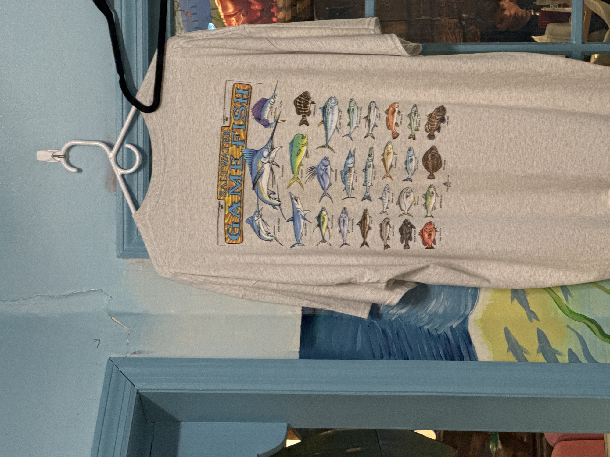 A gray t-shirt with a graphic featuring various saltwater fish is displayed on a white hanger against a light blue wall in Garden City Beach, United States of America. The graphic includes text "Saltwater Gamefish." The wall has an ocean-themed painting with fish and waves at the lower portion of the frame. Part of a dark-colored hanger can be seen. Some reflections and other objects are vaguely visible through a window.