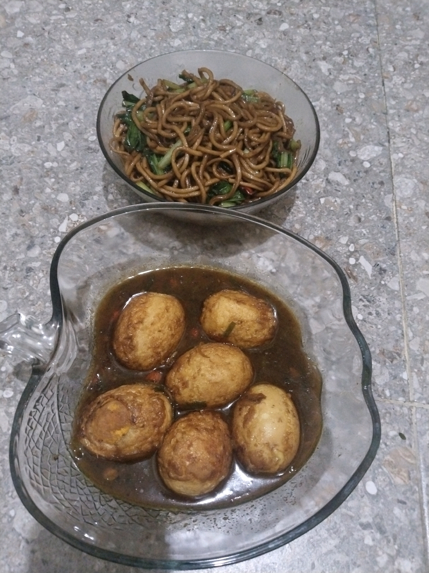 The image shows two bowls of food placed on a textured gray surface. The upper bowl contains noodles and some green vegetables, likely stir-fried. The lower bowl is made of clear, patterned glass and holds several whole eggs submerged in a dark, savory sauce. The eggs appear to be hard-boiled and possibly braised or stewed. The overall presentation suggests a home-cooked meal. The location is described as Kuripansari, Indonesia.