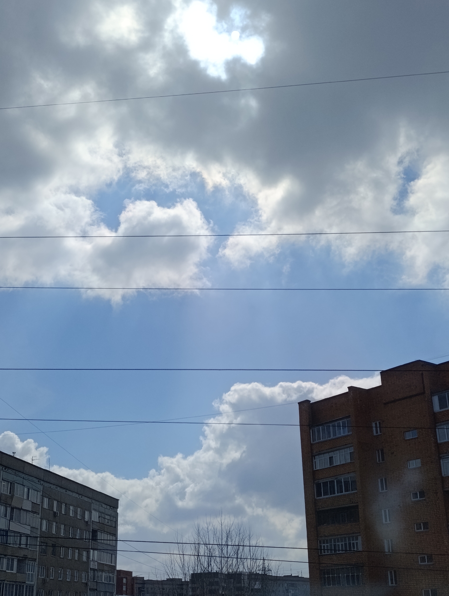This upward-facing photograph captures a partial view of an urban sky dominated by clouds and glimpses of residential buildings. The upper two-thirds of the image are filled with a dynamic sky, featuring both bright white, fluffy cumulus clouds and darker, denser stratocumulus clouds. Patches of clear blue sky are visible, especially towards the center and lower left, where the sun's bright glare peeks through the clouds.
Multiple thin, dark power lines or utility cables stretch horizontally and diagonally across the frame, segmenting the sky and passing in front of the buildings.
In the lower portion of the image, the tops of two distinct multi-story residential buildings are visible. On the left, a grey, concrete-paneled building with numerous square windows and balconies suggests a typical post-Soviet architectural style. On the right, a taller, reddish-brown brick building stands prominently, featuring many large windows, some with enclosed loggias or balconies, indicating a more modern construction or facade. Bare, leafless trees are visible in the lower-center, further suggesting late winter or early spring in a temperate climate.
The scene depicts an ordinary daylight moment in Krasnoyarsk, Russia, characterized by the specific architectural styles common to the region and the visible utility infrastructure. There are no people or specific activities taking place, and no discernible text is visible on the buildings or elsewhere.