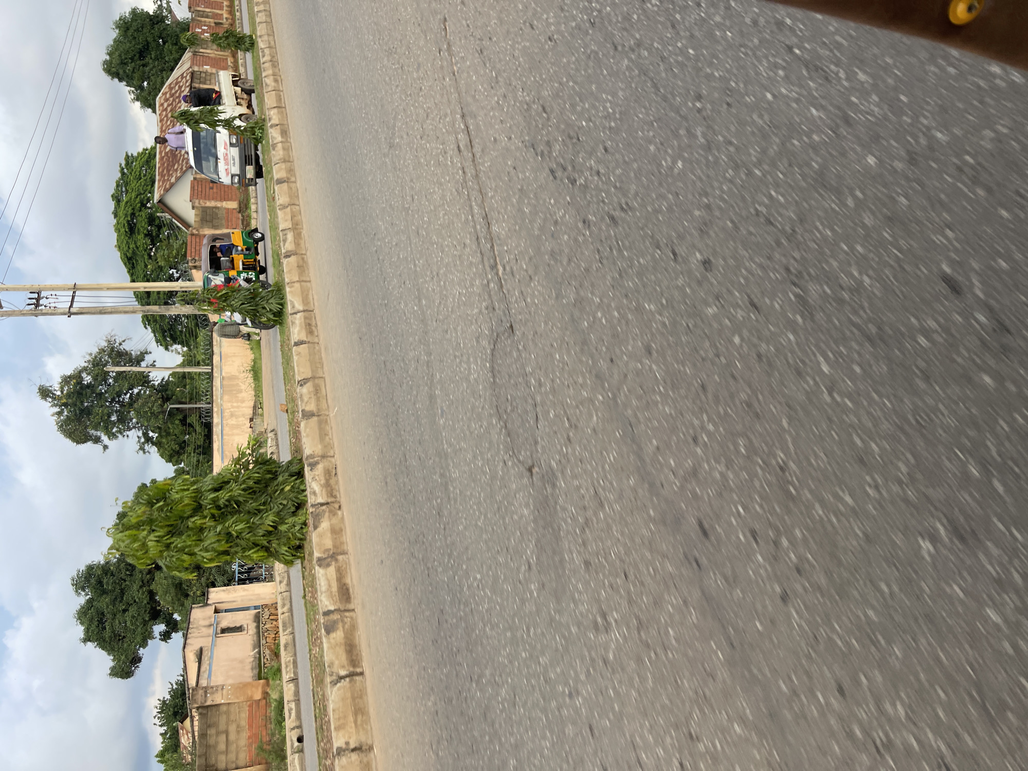 The image depicts a roadside scene in Jos, Nigeria. In the foreground, a paved road with visible cracks and an uneven surface suggests it is a well-trafficked area. The road is bordered by a concrete curb and a strip of grass.
Further back, a light-colored truck with a flatbed is parked on the side of the road. Two individuals are visible on the truck bed, one standing and wearing a light purple shirt, and another seated. Next to the truck is a three-wheeled autorickshaw, commonly known as a Keke Napep, with a driver and a passenger. In the distance, buildings with beige walls and a corrugated iron roof are visible, along with a few trees. A utility pole with electrical wires stands prominently. The sky is overcast with clouds. The overall impression is a typical urban street in Nigeria, characterized by its modes of transportation and architecture.