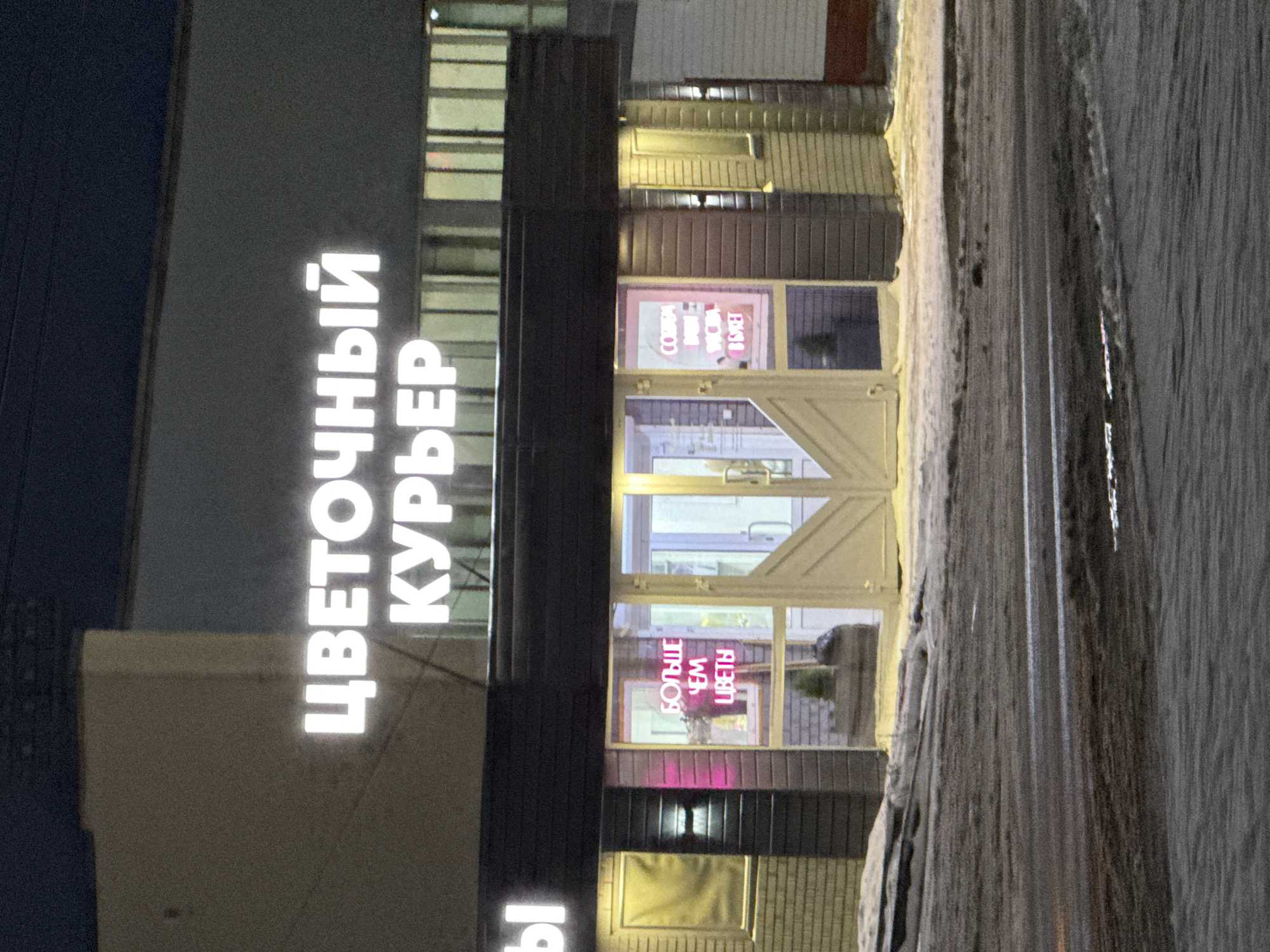 The image shows a flower shop at night. The sign reads "Цветочный Курьер", which translates to "Flower Courier". The shop is located in Votkinsk, Russia. Neon signs in the windows read "Собираем Душой Дарим В Свет" and "Больше чем цветы", meaning "We collect with soul, we give in light" and "More than flowers." The street is wet. No people are visible.