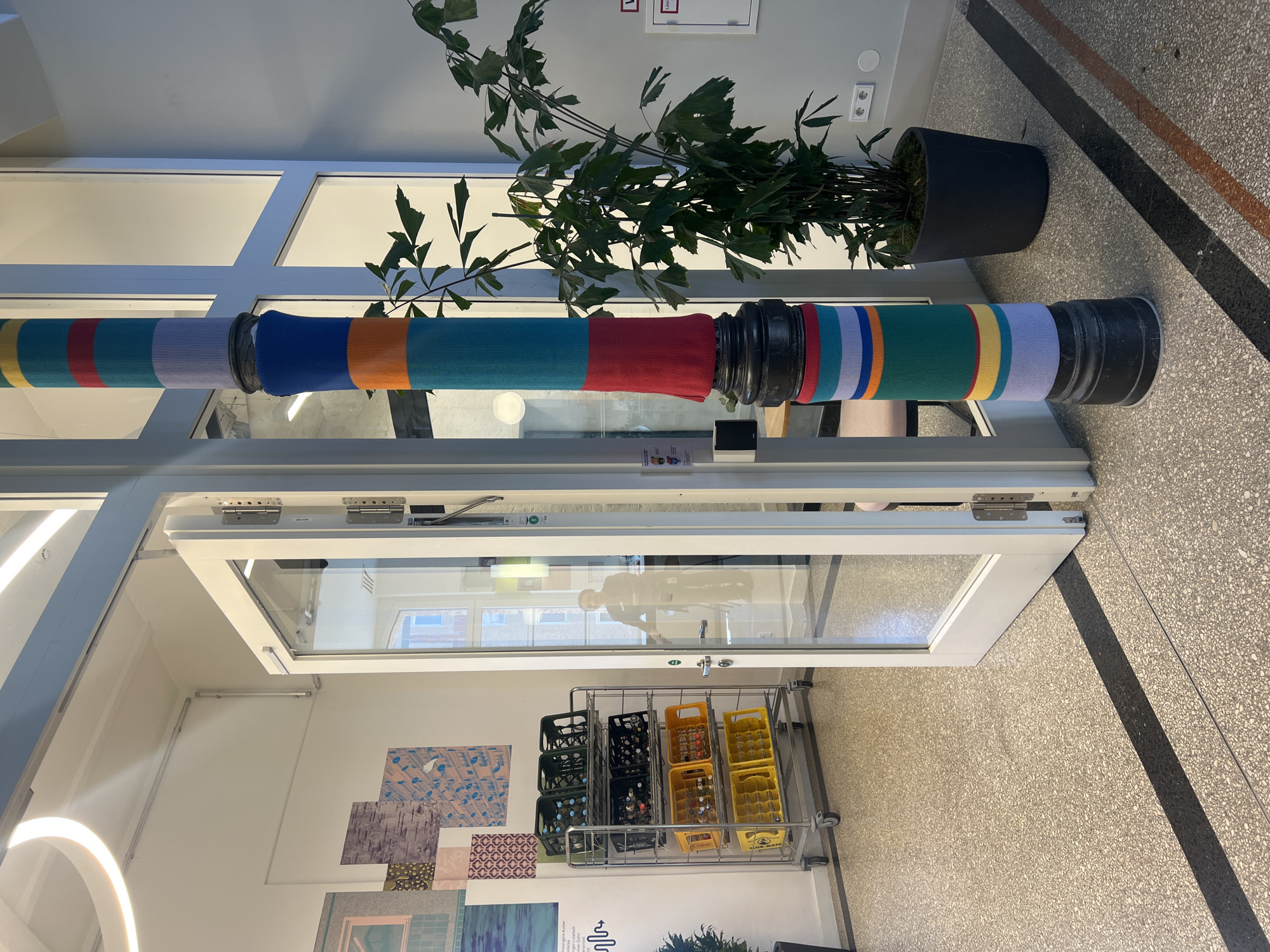 This image depicts an indoor scene, likely a hallway or communal area within a modern building, potentially in Berlin, Germany. The central feature is a large, horizontal pipe or beam, spanning the upper-middle section of the frame, which is decoratively wrapped in several sections of colorful, horizontally striped fabric or knitted material. These "yarn-bombed" segments display vibrant hues including blue, green, red, orange, yellow, and light purple, and are separated by black, industrial-style connectors.
To the left, positioned behind the decorated pipe, a large dark green plant with broad leaves sits in a black pot on the floor. The floor itself is a light-colored, speckled terrazzo or concrete, marked by a distinct diagonal dark gray stripe.
On the right, a white-framed glass door stands partially ajar, revealing a reflection of an interior space with another window and a faint, elongated human reflection. Below this open door, a metal rolling cart is filled with various plastic bottle crates. Yellow crates are visible, some containing bottles, with the discernible text "COKE" and "KLAMMATH." Green and black crates, also holding bottles, are present on the cart.
The white wall behind the cart is adorned with multiple squares of patterned, abstract artwork. The space is brightly lit, suggesting daytime. The scene is static, showcasing a decorated and functional interior.