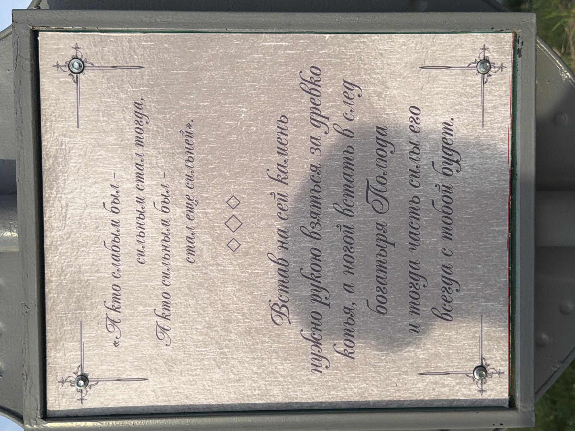 This image shows a weathered plaque mounted on a dark grey metal post in Cherdyn, Russia. The plaque is rectangular with ornate borders and four decorative corner elements, each containing a small screw. The text on the plaque is in Cyrillic script and appears to be an excerpt from a poem or proverb. The lighting suggests it is daytime, with shadows cast on the plaque from the post. The background is slightly out of focus but appears to be an outdoor setting with hints of green foliage. The plaque itself has a textured, metallic finish that shows some signs of wear and tear.