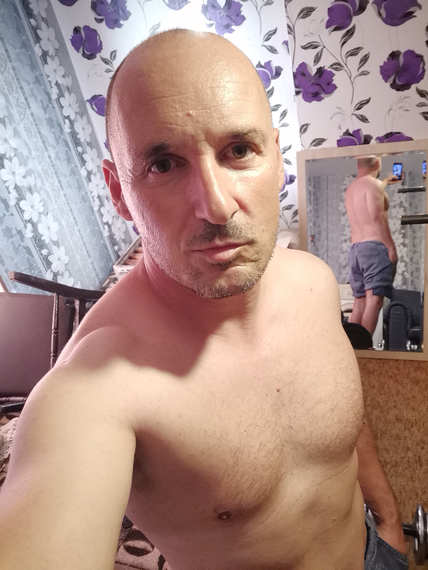 The image captures a selfie of a bald man with a serious expression, his face and scalp appearing sweaty. He has short facial stubble and is shirtless, revealing a hairy chest and shoulders.
In the background to the right, a mirror reflects what appears to be the same man, viewed from behind. He is shirtless and wearing blue shorts, holding a smartphone, suggesting he is also taking a picture or recording himself. Partially visible exercise equipment and a dumbbell on the floor further support the idea of a workout setting.
The room features wallpaper with a white background and prominent purple floral patterns. A sheer white curtain with a white floral design hangs on the left side of the frame. Part of a wooden chair is visible behind the main subject on the left. The lower section of the wall or floor has wooden paneling.
The overall scene suggests an indoor setting, likely a home gym or personal workout space, with the man documenting his physical state after exertion. There is no visible text in the image.