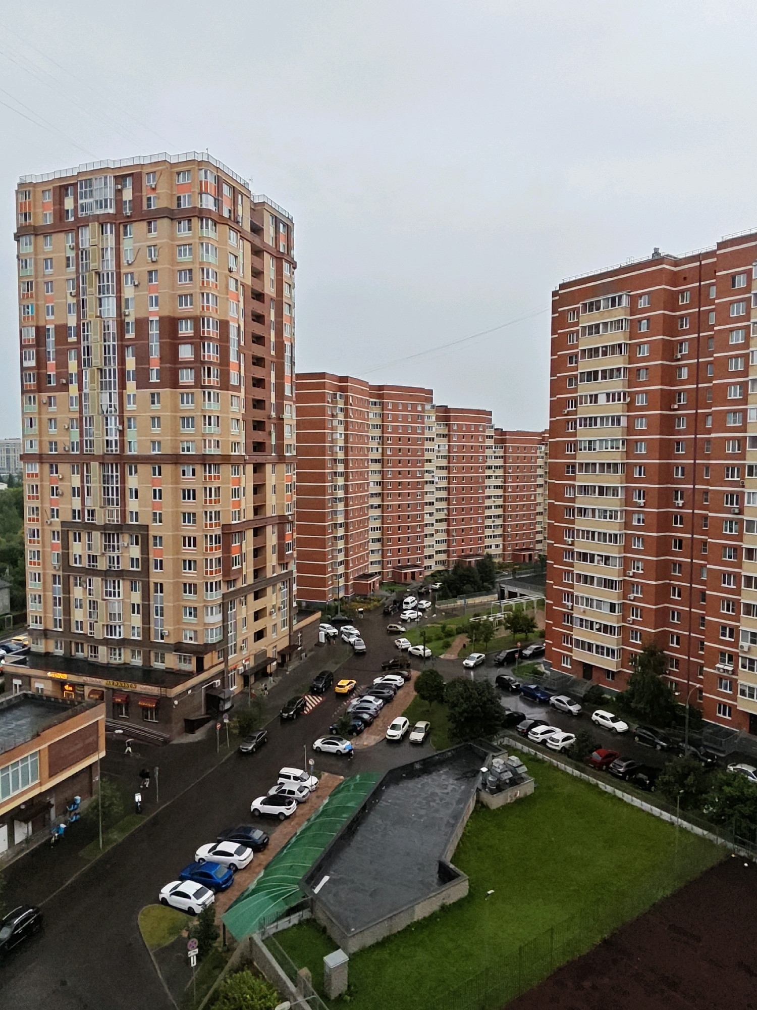 This is an aerial, eye-level, medium shot of a residential area in Shcherbinka, Russia, on an overcast day. The scene is dominated by several tall, multi-story apartment buildings. The building on the left is a light brown and terracotta color with a complex facade featuring many windows and balconies. To the right, a series of identical, brick-red apartment buildings with white trim stretches into the distance. A street runs between the buildings, lined with parked cars. Some of the cars are white, silver, black, and one is yellow. There are also sidewalks and patches of green grass and trees. A dark, flat-roofed building is in the foreground on the lower left. Two people are visible on the sidewalk near the dark building, and a few more are further down the street. The overall impression is of a typical urban residential environment.