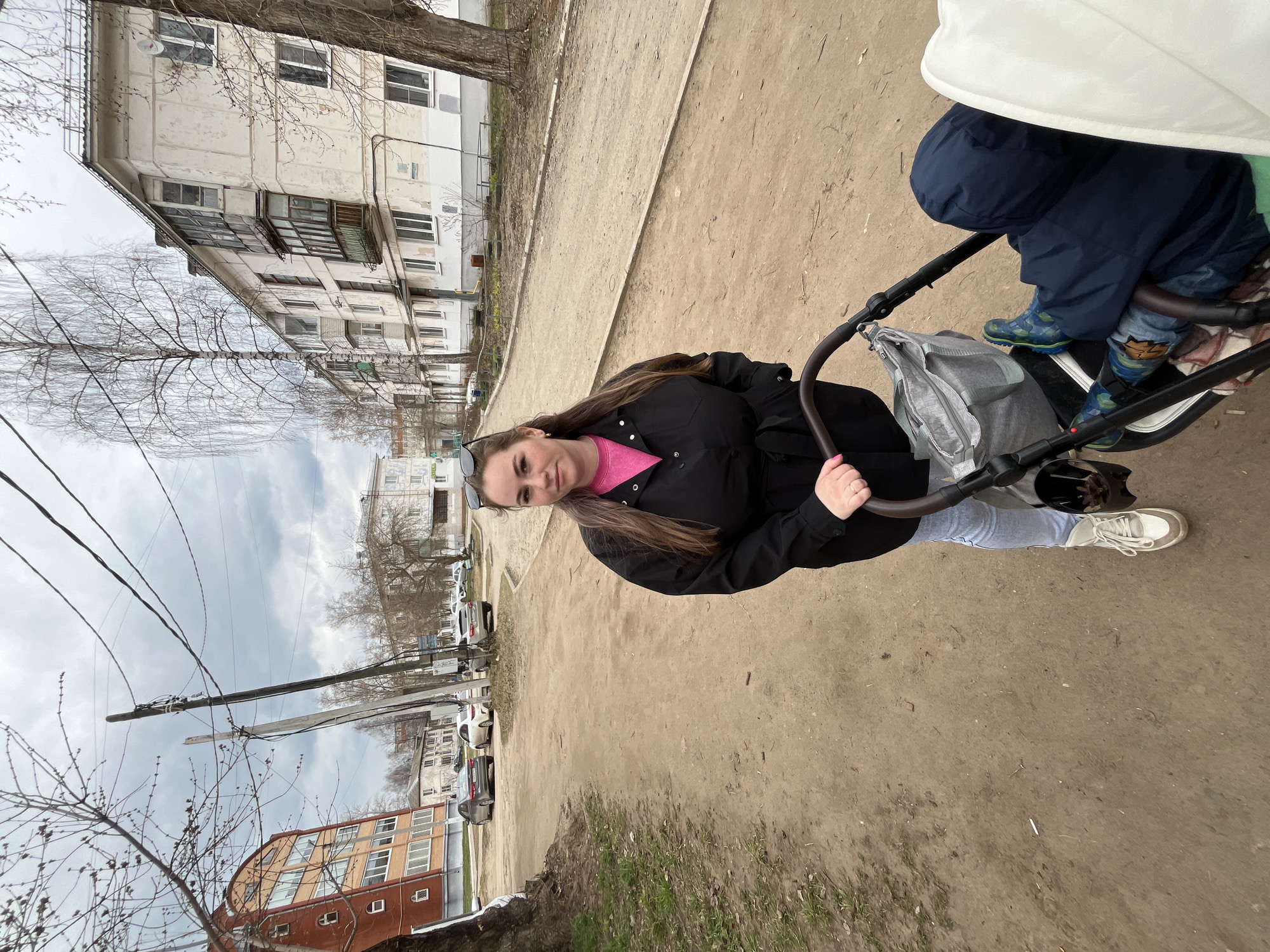 The image captures a woman pushing a stroller on a dirt path in an urban or suburban residential area, likely in Chaikovsky, Russia. The sky is overcast, and the leafless trees suggest late winter or early spring.
The main subject is a woman, appearing to be in her 20s or 30s, with long brown hair. She is dressed in a black jacket over a pink top, light blue jeans, and white sneakers. Sunglasses are resting on her head. She looks directly at the camera with a slight, calm smile while holding the handle of a dark-framed stroller. A grey bag is attached to the stroller's handle.
Within the stroller, a child is partially visible, wearing a dark blue jacket and distinctive patterned boots. The stroller has a light-colored canopy.
In the background, several multi-story apartment buildings are present. To the left, a light-colored, slightly worn building with visible windows and balconies stands. Further along the path, a reddish-brown building with many windows can be seen. A utility pole with numerous power lines extends across the frame, and several parked cars are visible in the distance. The ground is a mix of dirt and gravel, with occasional patches of sparse grass. A white cigarette butt lies on the ground near the woman's feet. The scene portrays a casual daytime outing with a child in a residential neighborhood. No visible text is present.