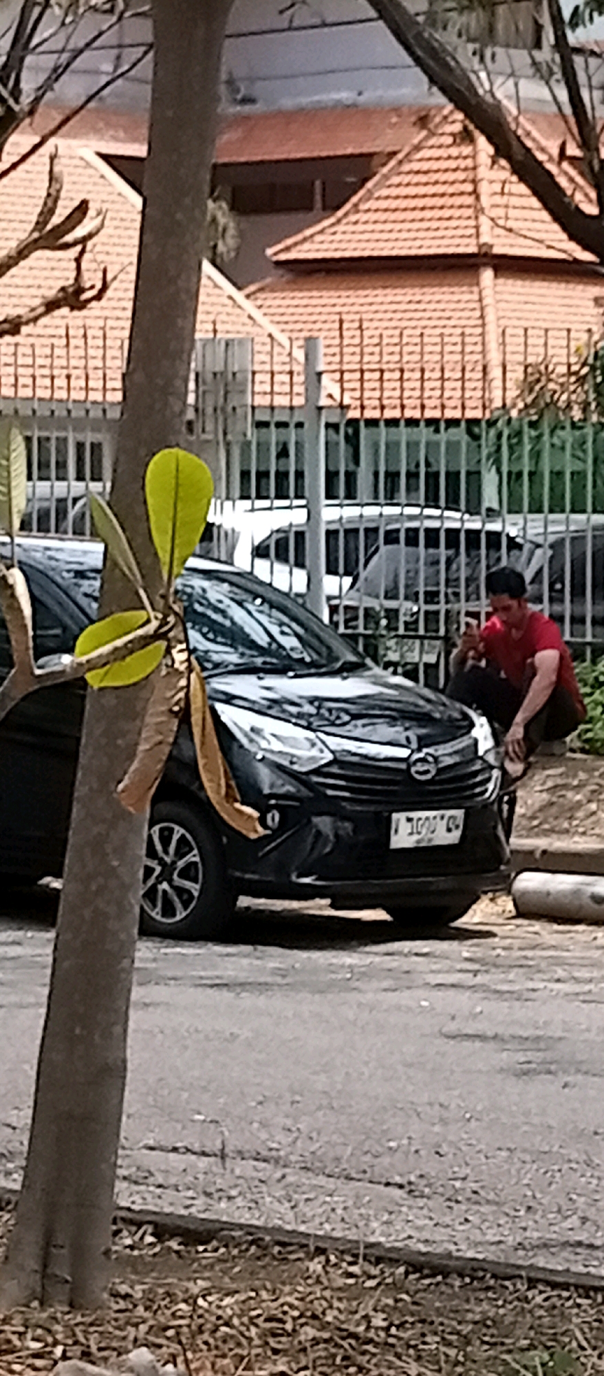 A man in a red shirt is crouching next to a black car in what appears to be a residential area in Surabaya, Indonesia. He is holding a mobile phone. The car is parked on a paved surface, possibly a street or driveway, with a concrete barrier in the foreground. In the background, there is a fence and buildings with tiled roofs. A tree trunk with green leaves and some brown fabric draped around it is in the foreground, partially obscuring the car. The overall scene suggests an urban or suburban environment.