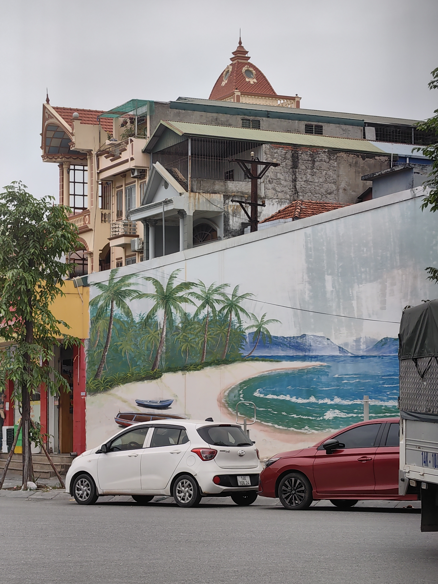 The image captures a vibrant street scene in Viet Hung Ward, Vietnam, under an overcast sky. In the foreground, a long white wall dominates the view, adorned with a large, colorful mural of a tropical beach. The mural depicts a sandy crescent beach lined with numerous green palm trees, meeting clear blue ocean water with white waves. Distant mountains are visible across the water, under a light blue sky. Two small boats, one blue and one reddish-brown, are painted on the sand.
Parked on the asphalt street directly in front of the mural are two cars. A white Hyundai i10 hatchback is prominently positioned in the center, displaying the Vietnamese license plate "14K 106 24". Behind it, a red sedan, possibly a Honda City, is partially visible. On the far right, a large dark green or grey truck is partly seen, with the text "14K-10" visible on its side.
Behind the mural and the parked vehicles, a dense array of buildings rises. On the left, a distinctive multi-story building features an ornate, reddish-tiled roof with a pointed spire and decorative arched windows and balconies. Adjacent to it, another building has a rooftop area covered by a green mesh structure. Further back and to the right, a building with a prominent red-domed roof, adorned with circular windows, stands out. Various other residential or commercial structures, some with corrugated metal roofs and barred windows, complete the dense urban background. A utility pole with multiple cross arms is visible against the sky, positioned between the buildings. A small tree with green leaves is on the left, next to a yellow-painted building facade. The scene suggests a calm, everyday urban environment.