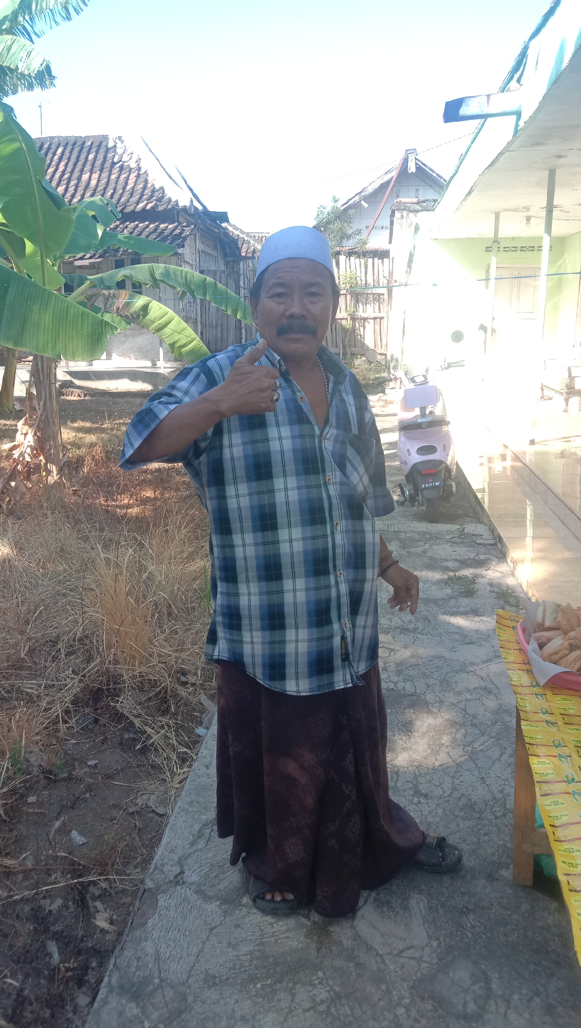 In Ngempit, Indonesia, a man with a mustache and wearing a blue cap and a blue and white plaid shirt gives a thumbs-up. He is standing on a concrete path in front of a modest building with a green wall and a light-colored roof. To his left are banana trees and some dry grass. To his right, a pink scooter is parked. A wooden table with food items is also visible on the right. The sky is clear and bright.