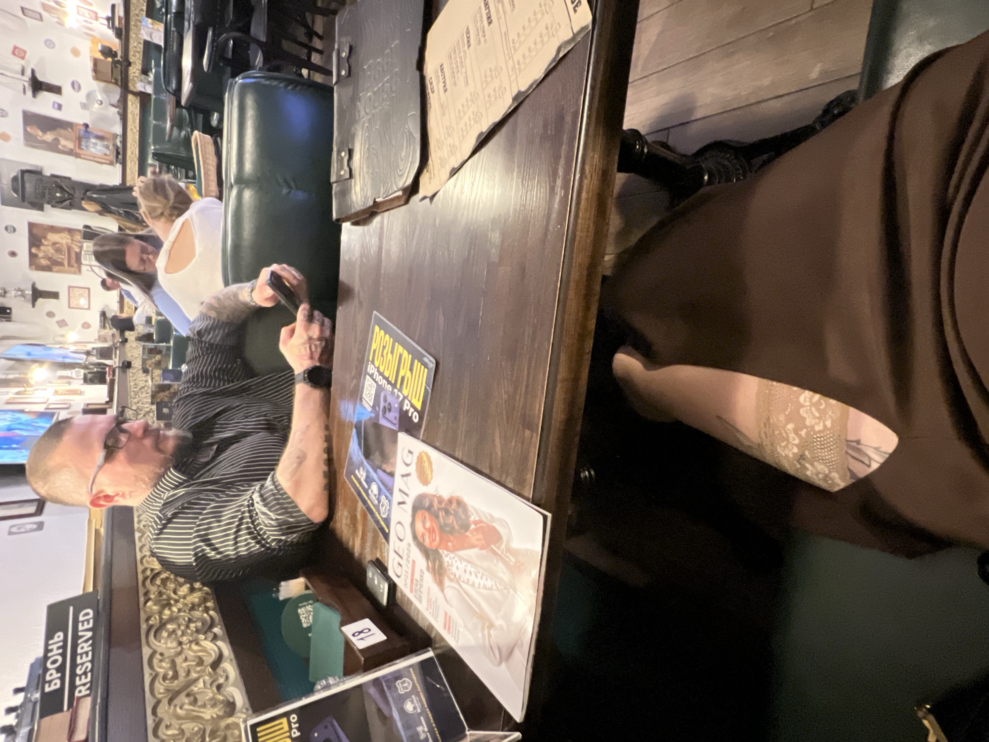 The image depicts an indoor scene in what appears to be a restaurant or cafe setting in Saint Petersburg, Russia. A middle-aged man with glasses and a striped shirt is seated at a wooden table. He appears to be interacting with a cell phone. On the table, there are several magazines, including one titled "GEO MAG." There is also a reserved sign in Russian and English, "БРОНЬ RESERVED". To the right, there is a person wearing a brown dress sitting in the same table. Other people are visible in the background, indicating a populated environment. The restaurant has ornate, classical decor.