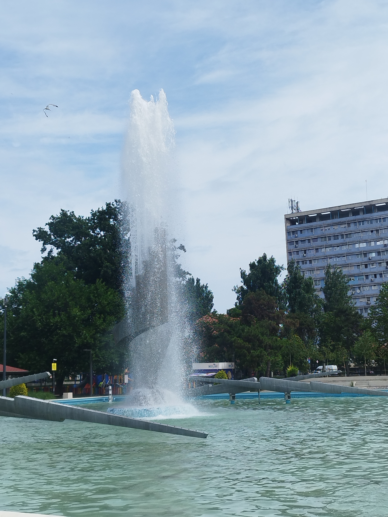 This image captures a public square with a large, multi-tiered fountain as its centerpiece. The fountain is actively spraying water high into the air, creating a dynamic visual effect against the sky. The water is a light, almost ethereal white, contrasting with the darker, more structured elements of the fountain's base.
Surrounding the fountain is a pool of calm, turquoise water, reflecting the light and the sky. The edge of the pool features modern, angular concrete structures that seem to be part of the fountain's design, possibly serving as seating or decorative elements.
In the background, a cluster of lush green trees provides a natural element, softening the urban landscape. Behind the trees, a tall, grey, multi-story building dominates the skyline. This building has numerous windows and appears to be an apartment complex or office building, with visible antennas on its rooftop.
The sky is partly cloudy, with streaks of white clouds scattered across a light blue expanse, suggesting it is daytime, likely late morning or afternoon. The overall mood is serene and public, typical of a city park or plaza designed for recreation and relaxation. There are no people clearly visible in the frame, but the presence of playground equipment in the background suggests it's a family-friendly area. No text is clearly legible in the image. The scene is set in Constanța, Romania.