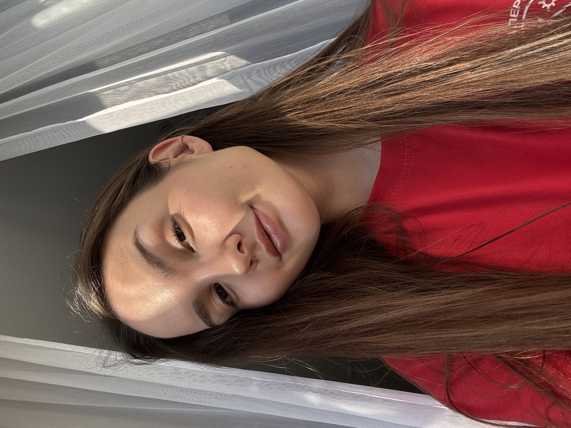 A young woman with long brown hair is lying down, her head tilted, in what appears to be an indoor setting near a window covered with a sheer white curtain. She is wearing a red t-shirt. Based on the text visible on the shirt, the location might be Chaikovsky, Russia. The room seems to be illuminated with natural light.