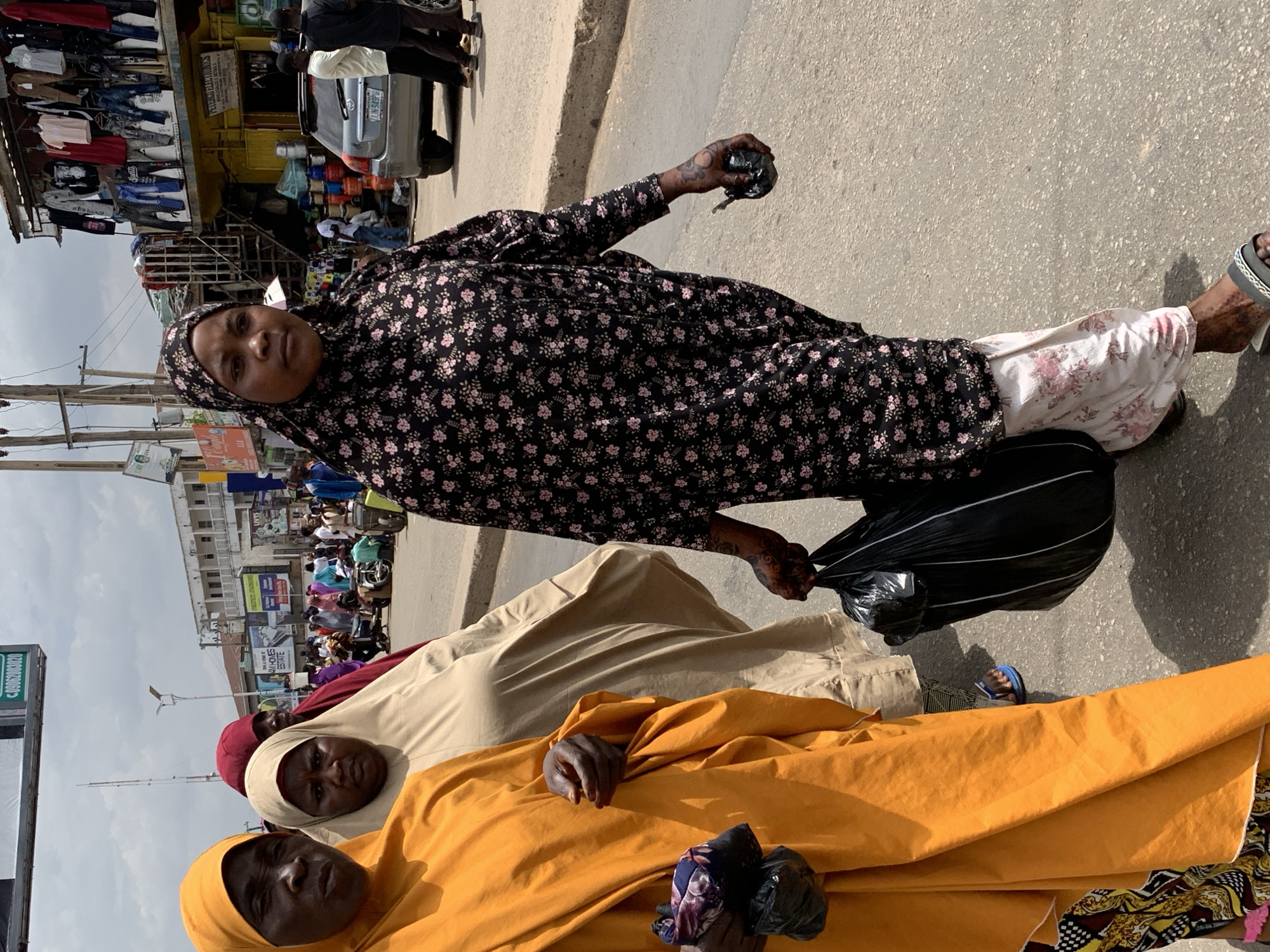 The image, rotated sideways, depicts a street scene in Jos, Nigeria. Three women wearing hijabs are visible. One is dressed in a black floral-patterned hijab and gown, carrying a black plastic bag. Another is draped in a tan/beige hijab and gown, and a third is wearing a bright yellow hijab and gown, also holding a dark object wrapped in plastic. The women are standing on a paved surface, likely a sidewalk. Behind them is a street with pedestrians, vehicles, and buildings. On the left, a building with clothing on display is visible. A silver car is parked nearby. The sky is overcast. The overall impression is that the women are in a busy urban area.