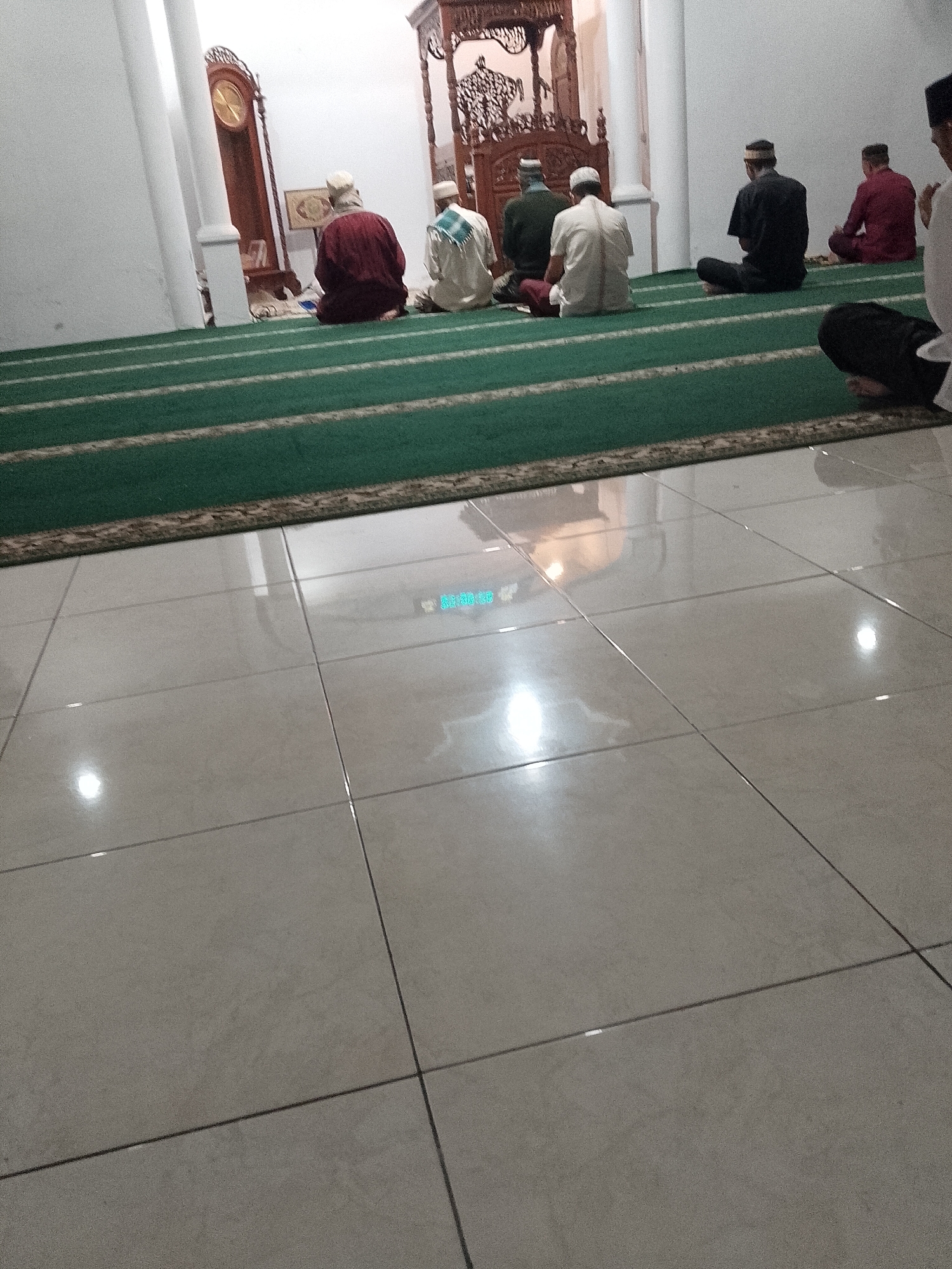 In a mosque in Ranoyapo, Indonesia, several men are seated on a green carpeted floor, facing away from the camera. They are engaged in prayer. To the left, a grandfather clock stands tall. The floor is tiled and reflects the overhead lighting. The walls are white.