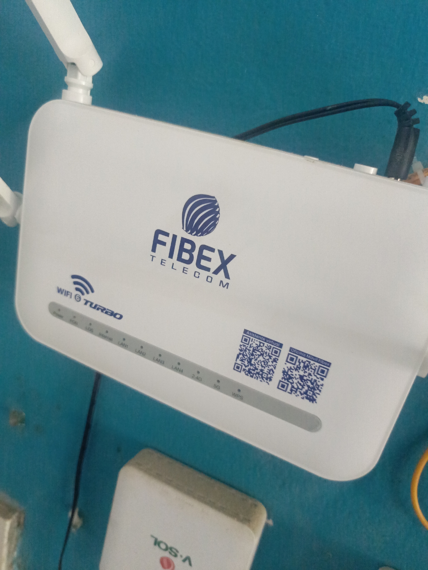 The image shows a FIBEX TELECOM WiFi router mounted on a turquoise wall. The router has two antennas and displays various indicator lights labeled Power, PON, LOS, Internet, LAN1-4, 2.4G, 5G, and WPS. QR codes labeled "Asistente virtual" and "Oficina Movil Fibex" are printed on the router's surface. The device is labeled "WIFI TURBO." A partial view of a power adapter is visible below the router, labeled "TOS A." A black cable connects to the router's back. The image does not show any indications that it's located in Juan Griego, Venezuela.