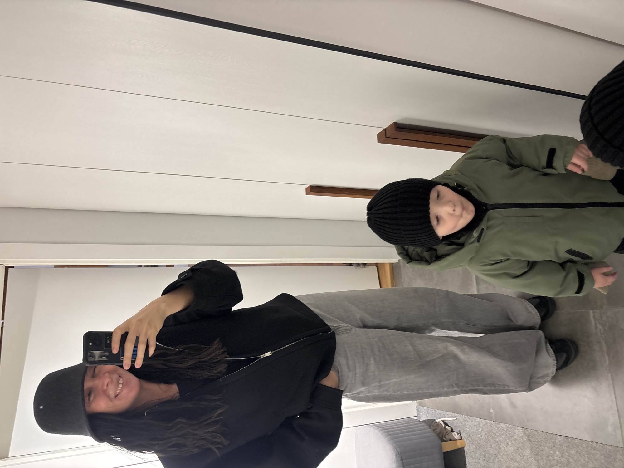 A woman and child are reflected in a mirror. The woman is wearing a black bucket hat, a black zip-up hoodie, and grey sweatpants. She is holding a phone, presumably taking the picture. The child is wearing a black beanie and a green jacket. They appear to be in a room with white walls and a light-colored floor. There's a glimpse of seating furniture in the bottom left corner. The location is unknown but possibly in Chaikovsky, Russia.