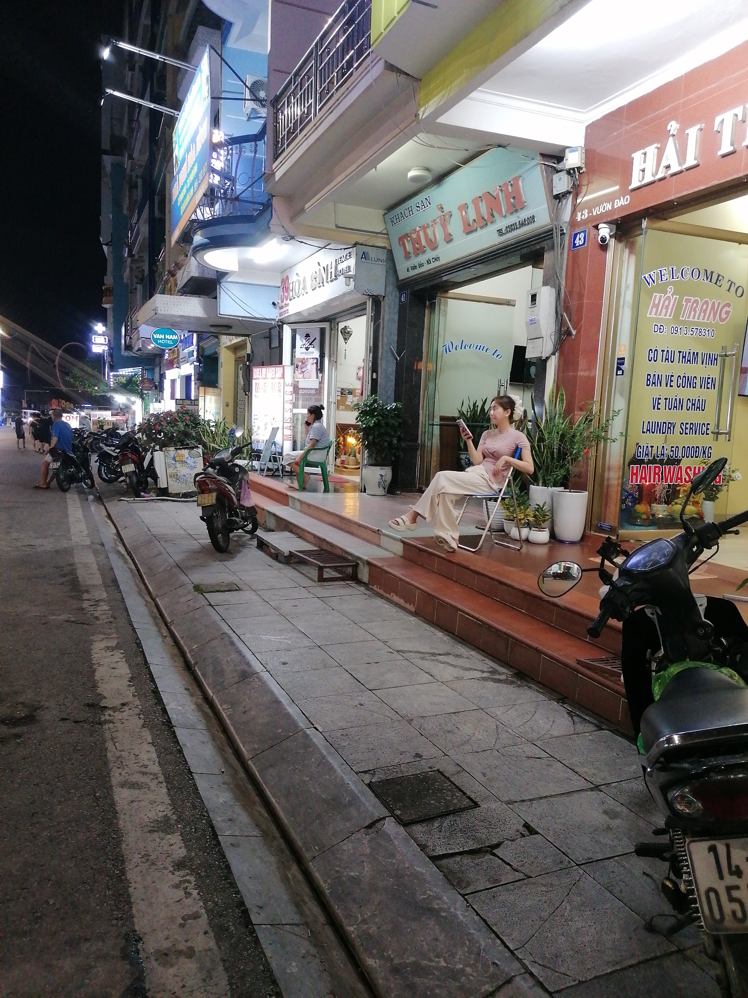 This nighttime street scene captures a vibrant commercial area in Bai Chay Ward, Vietnam. The image is dominated by the artificial glow of illuminated signs and building interiors, contrasting with the dark sky.
In the foreground, a concrete sidewalk lines an asphalt road with a faded white dividing line. Numerous motorcycles are parked along the curb, indicating a common mode of transport in the region. One motorcycle partially obstructs the view on the far right.
The central subject is a woman seated casually on a folding chair on a raised, tiled stoop outside a series of storefronts. She is dressed in a light-colored top and wide-leg trousers, holding a smartphone and looking towards the right side of the frame. Potted plants flank her on the stoop.
The surrounding buildings feature multiple businesses. Above the woman, a blue sign reads "KHÁCH SẠN THỦY LINH" (Thuy Linh Hotel), with an address "41 Viên Đào - BÃI CHÁY" and a phone number "TEL:02033.646.006". To her right, a red and white sign displays "HẢI TRANG" (Hai Trang), advertising services like "WELCOME TO HẢI TRANG," "CÓ TÀU THĂM VỊNH" (boat trip to visit the bay), "BÁN VÉ CÔNG VIÊN" (sell park tickets), "VỀ TUẦN CHÂU" (to Tuan Chau), "LAUNDRY SERVICE," and "GIẶT LÀ: 50,000Đ/KG" (laundry for 50,000 VND/kg), with "HAIR WASH" also visible. A "43" is visible on the pillar next to the Hải Trang entrance, which is at "43 - VƯỜN ĐÀO". A security camera is mounted above this entrance.
Further down the street, additional signs are visible, including "VAN NAM HOTEL" and "39 HÒA BÌNH HOTEL." People are scattered along the sidewalk, some near parked motorcycles, suggesting evening activity. Another person is seated on a green plastic chair on the sidewalk in the mid-ground. The overall atmosphere is bustling but relaxed, typical of an evening in a commercial district.