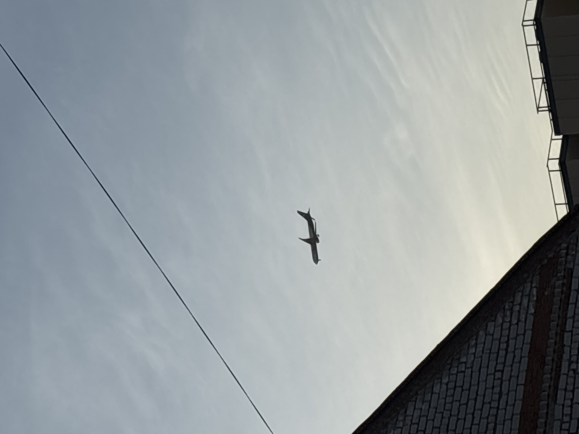 The image shows an airplane flying against a bright sky. A power line diagonally crosses the left side of the image. The corner of a brick building is visible in the lower right, and a section of a building with balconies is in the upper right. It is not possible to tell the location of Perm, Russia from just the image.