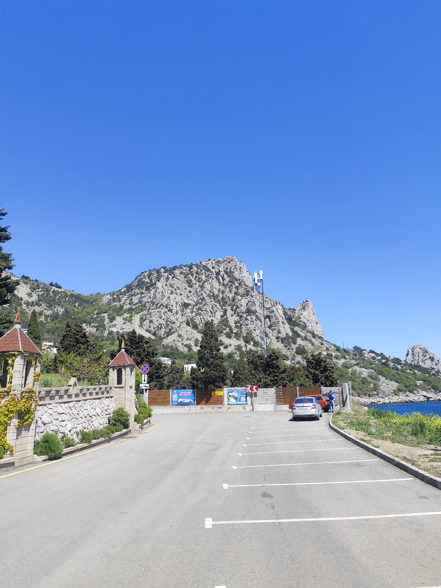 The image shows a parking lot with a mountainous backdrop under a clear blue sky. The parking lot is paved with white parking space lines. A car is parked towards the end of the parking lot near a concrete barrier separating the parking lot from an area of foliage and the sea. A person is standing near the car. Behind the parking lot, there is a low barrier with advertising posters. Behind the barrier, trees partially obscure a rocky mountain range. To the left of the parking lot, there is a decorative stone wall. The location is Unknown, Unknown.