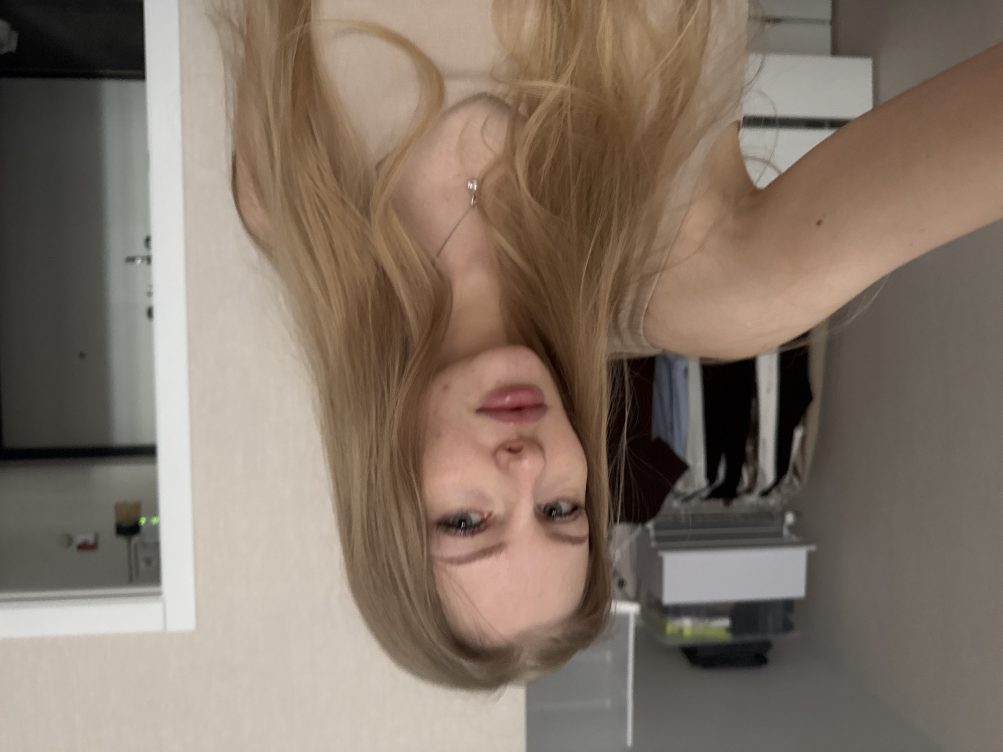 A young woman with blonde hair is taking a selfie indoors. She is facing the camera, and her hair covers part of her face. She is wearing a necklace. In the background, there is a cabinet or closet with clothes hanging inside. Also in the background are some boxes, a door and part of a wall, so the environment is likely a bedroom or apartment. There is no evidence to suggest this image was taken in Perm, Russia, or anywhere else. The location is unknown.