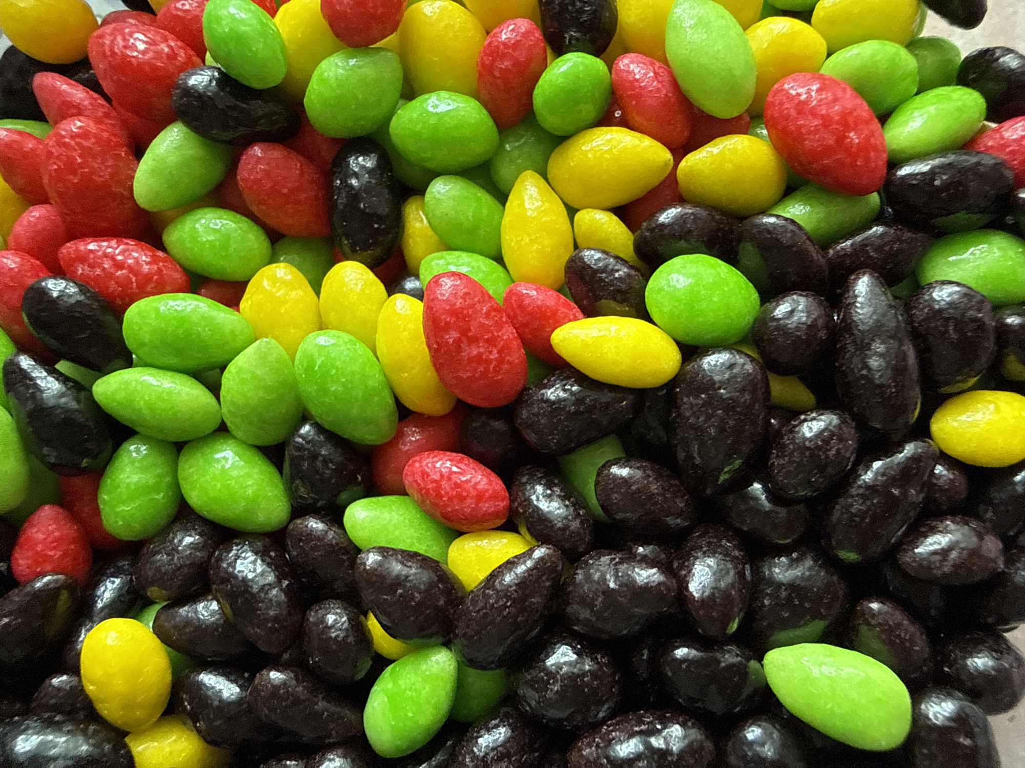 The image is a close-up, overhead view of a pile of colorful candies. The candies are oval-shaped and appear to be coated in a glossy shell. They are predominantly dark brown, bright red, lime green, and bright yellow. The candies are tightly packed together, obscuring any background. There is no discernible location in the city of Chaikovsky, Russia, or any people present in the image.