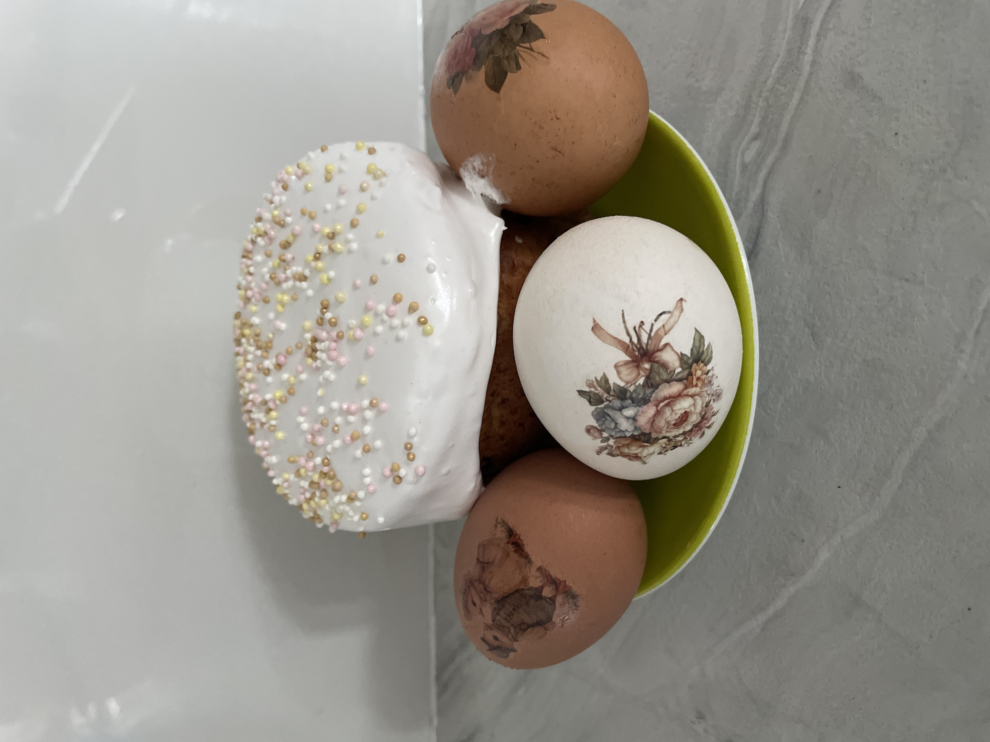 The image shows a green bowl containing an Easter cake decorated with white icing and sprinkles, along with three decorated eggs. The eggs feature floral designs. The scene is likely indoors, situated on a countertop with a light-colored surface, but there is not enough information to provide an exact location in the city of Лесной Пейзаж Кп (шарапово, Russia.