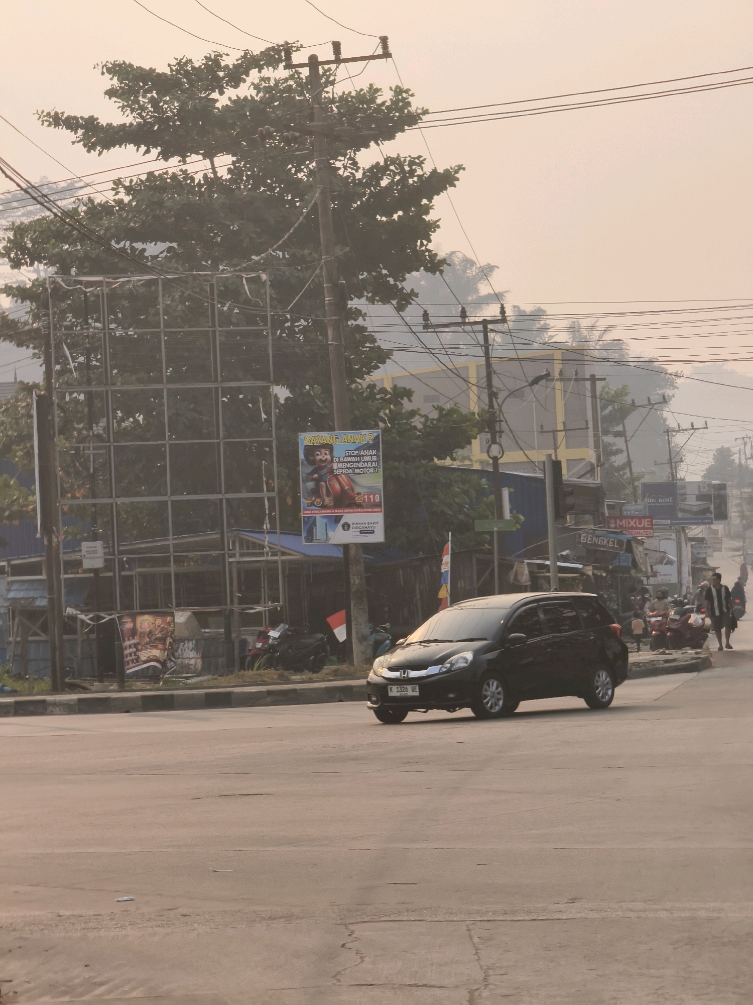 A black Honda Mobilio drives through an intersection in Samarinda, Indonesia, on a hazy day. The car's license plate reads "K 1326 VE". In the background, a billboard advertises for the hospital "RUMAH SAKIT DIRCAHAYU". Several people walk along the sidewalk, and motorcycles are parked in the background. Trees and power lines are visible throughout the scene. The sky is a pale orange, indicating potential smog or dust. The road is paved with asphalt and shows some wear and tear.