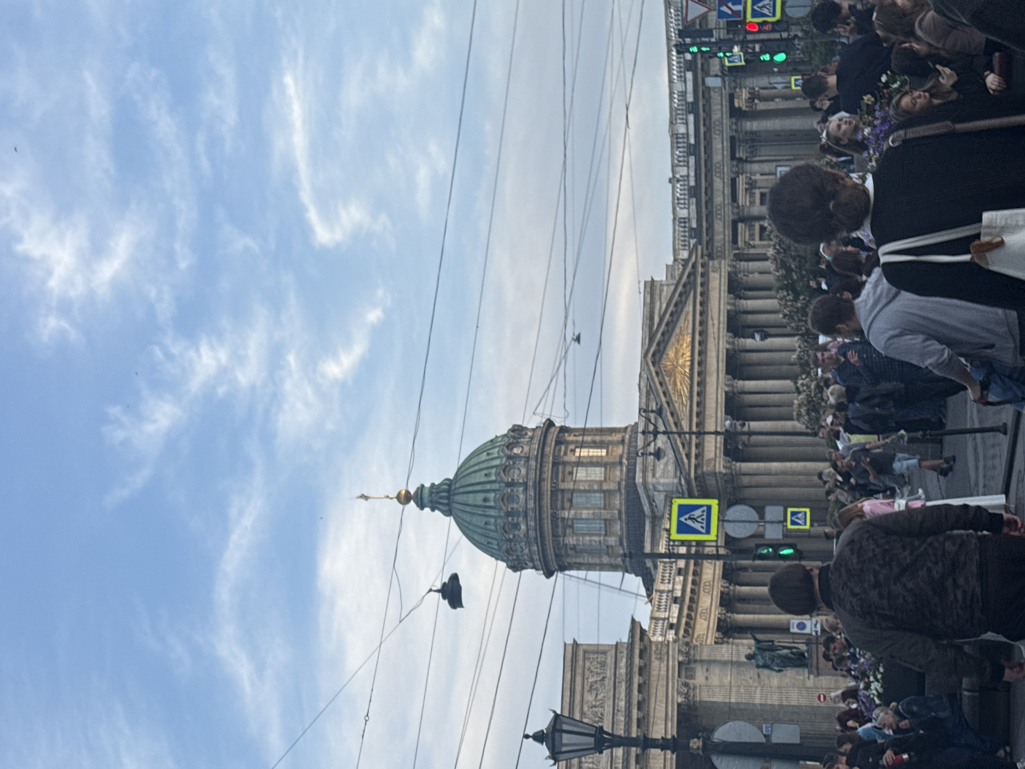 In Saint Petersburg, Russia, Kazan Cathedral is visible with its iconic green dome. The cathedral features classical architecture with a colonnade. A streetlamp stands to the left of the building. Multiple people are walking on the sidewalk, some gathered in groups. A pedestrian crossing signal displays a green light. Overhead, numerous wires crisscross the sky. The sky is partly cloudy with a light blue hue.