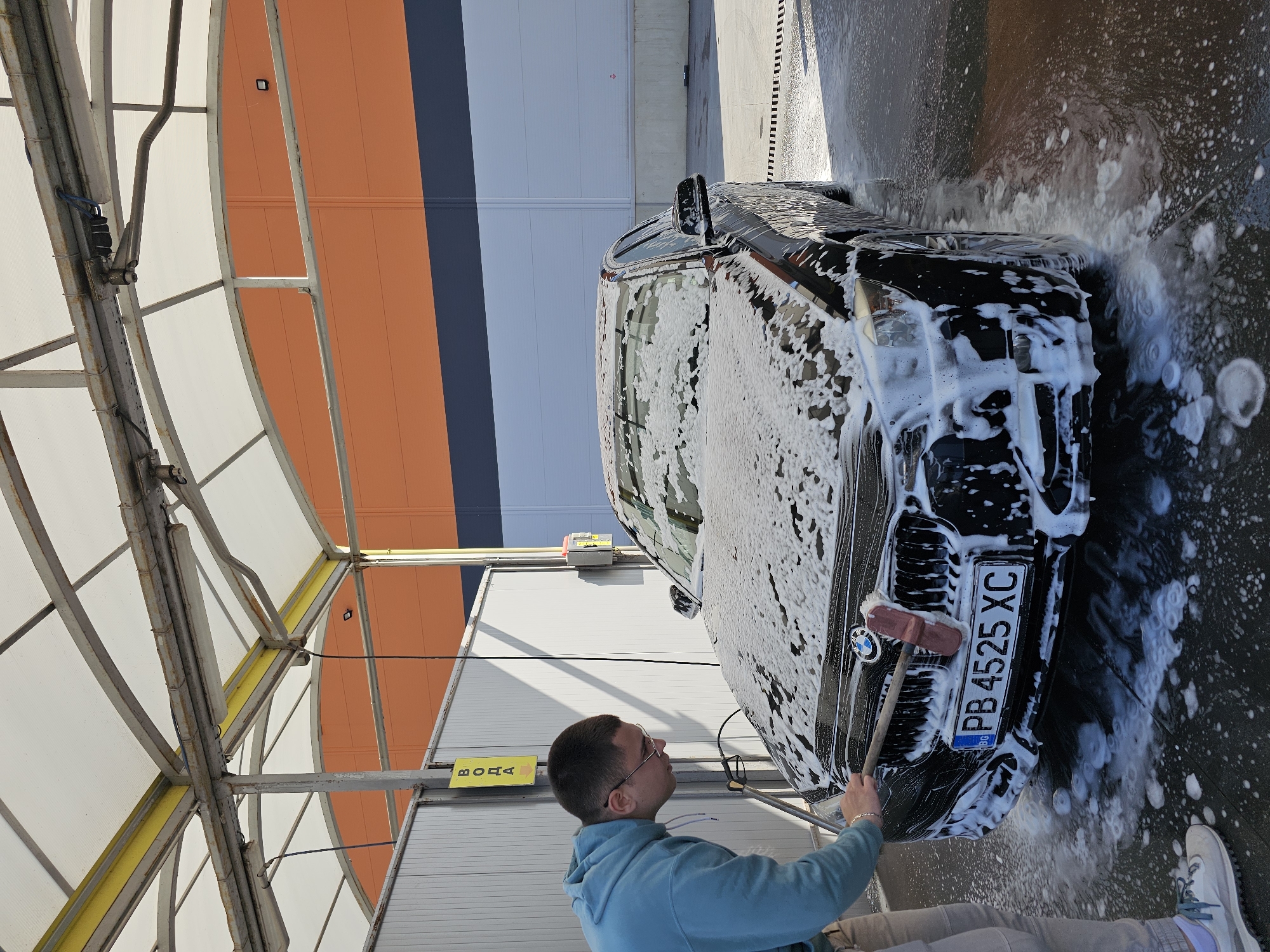 A man is washing a black BMW in a car wash bay. He is using a brush on a long handle to clean the front of the car, which is covered in white soapy foam. The car has a Bulgarian license plate that reads "PB 4525 XC." The man is wearing glasses and a light blue hoodie, and light-colored pants, and he has a bracelet on his left wrist.
The car wash bay has a metal frame and a semi-transparent roof. There is a sign with "ВОДА" (water) and an arrow on the wall. The background shows a building with orange, grey, and white stripes.
Based on the license plate (PB) and the context, the location is likely in or near Plovdiv, Bulgaria.