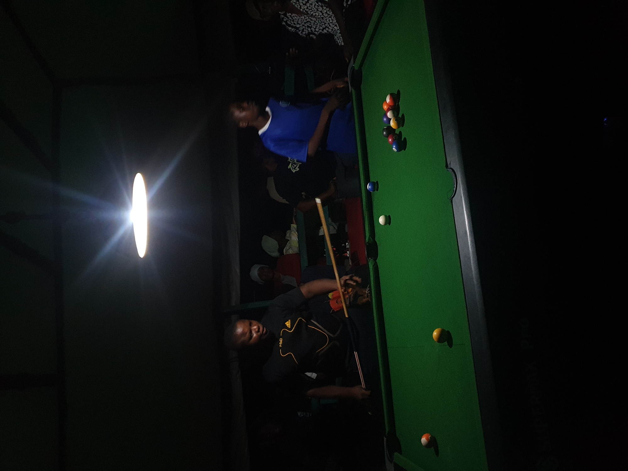 In a dimly lit room, likely a bar or recreational center in Lokoja, Nigeria, a group of individuals are gathered around a pool table. The scene is illuminated by a single, bright overhead light, creating strong shadows and highlighting the green felt of the pool table.
Two men are prominently featured. One, wearing a blue t-shirt, appears to be observing the game. The other, in a black t-shirt with yellow accents, is actively playing pool. He is in the process of taking a shot, holding a pool cue and focused on the balls on the table. Several pool balls are scattered across the green surface, including a rack of striped and solid balls near the top and a few individual balls in other positions.
Other people are partially visible in the background, suggesting a social gathering. The overall atmosphere seems casual and focused on the game. The lighting and the clothing suggest it is likely evening or night. There is no visible text in the image.