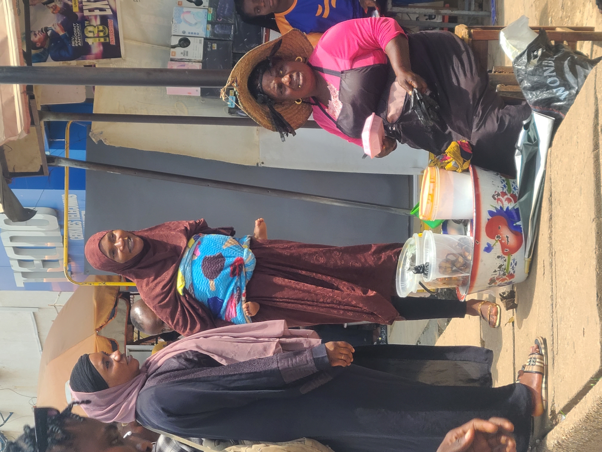 The image captures a street scene in Jos, Nigeria. In the foreground, a woman wearing a straw hat, a pink shirt, and a dark apron sits on a wooden stool, holding a pink plastic container. She has large gold hoop earrings and her expression suggests she is speaking or interacting with someone. Beside her, on the ground, is a patterned bowl filled with food items, as well as some plastic containers and bags.
To the left of the woman, two other individuals are visible. One is a woman in a dark abaya and a light pink hijab, facing to her right. Next to her, another woman is wearing a maroon hijab and a patterned cloth wrapped around her, with a baby visible underneath the cloth. The baby's feet are exposed.
The background reveals a street stall or shop with merchandise on shelves. A large blue and white sign with the word "TECNO" is prominent, indicating a telecommunications or electronics store. A poster featuring a man with a microphone and text related to an "Anniversary" is also visible. The ground appears to be paved with concrete, and the overall environment suggests a busy market or commercial area. The lighting indicates it is daytime.