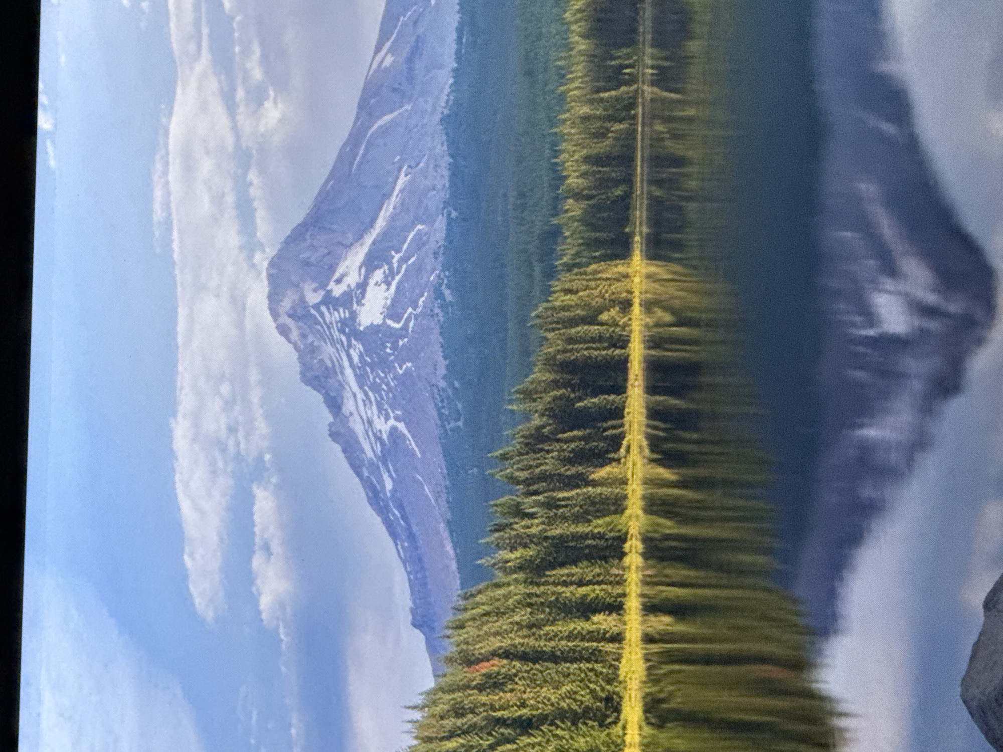 The image depicts a serene natural landscape featuring a snow-capped mountain, a dense evergreen forest, and a calm lake reflecting the scene. The mountain dominates the background, with white snow contrasting against its rocky slopes. Below the mountain, a thick line of dark green pine trees forms a distinct border. The foreground is occupied by a body of water, its surface smooth and mirroring the trees and mountain above. The sky is a light blue with scattered white clouds. The scene evokes a sense of tranquility and natural beauty. There is no indication that this scene is located in Saratov, Russia, or any other city. The landscape appears to be a natural wilderness setting.