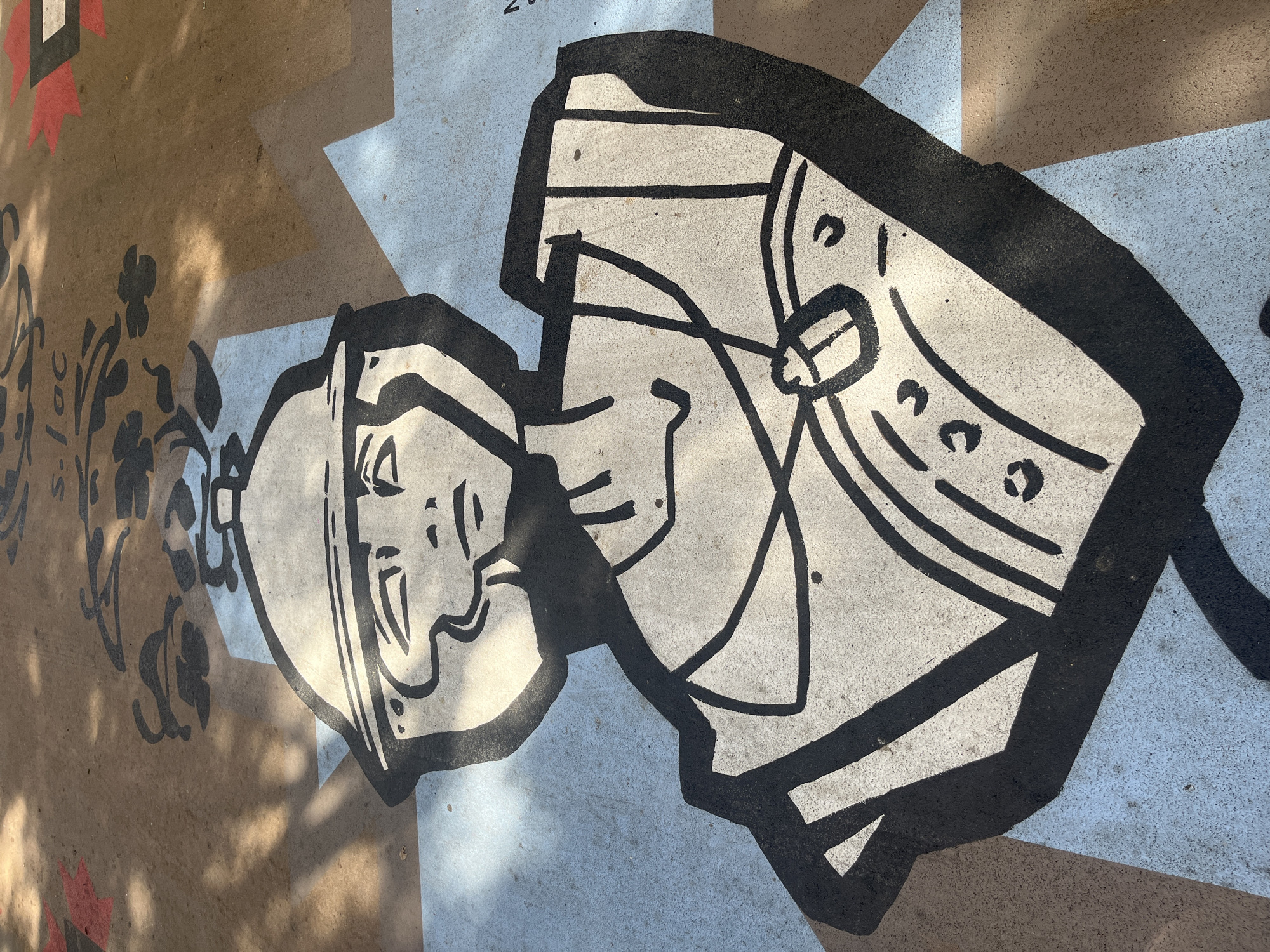 The image shows a stylized mural painted on the pavement in the city of Tarragona, Spain. The mural depicts the bust of a person wearing a helmet, with bold black lines defining the features and clothing. The person has a neutral expression, with closed eyes and a simple nose and mouth. They are wearing a helmet with a chin strap and a necklace with a pendant. The mural is set against a background of light blue and brown geometric shapes. Shadows from overhead trees are cast across the pavement, indicating an outdoor location. The pavement itself appears to be made of concrete or asphalt. The overall style of the artwork is graphic and illustrative.