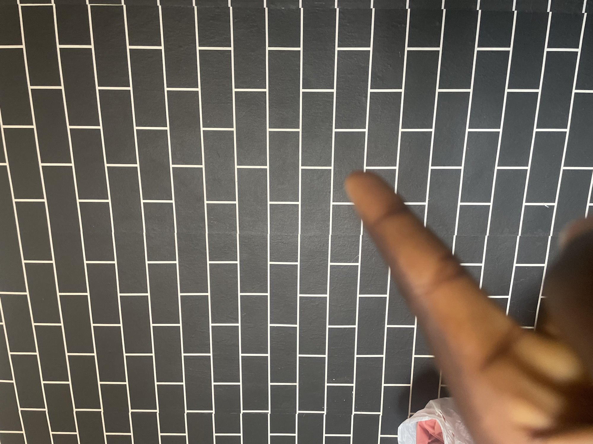 The image depicts a dark gray brick wall with white grout lines, arranged in a staggered pattern. A person's hand and finger, with medium-dark skin tone, are in the foreground, partially obscuring the wall and a white plastic bag containing pink items to the left. The scene is indoors, and the wall appears to be a feature of a building in Jalingo, Nigeria. The lighting is neutral, and the image is focused on the wall and the foreground elements.