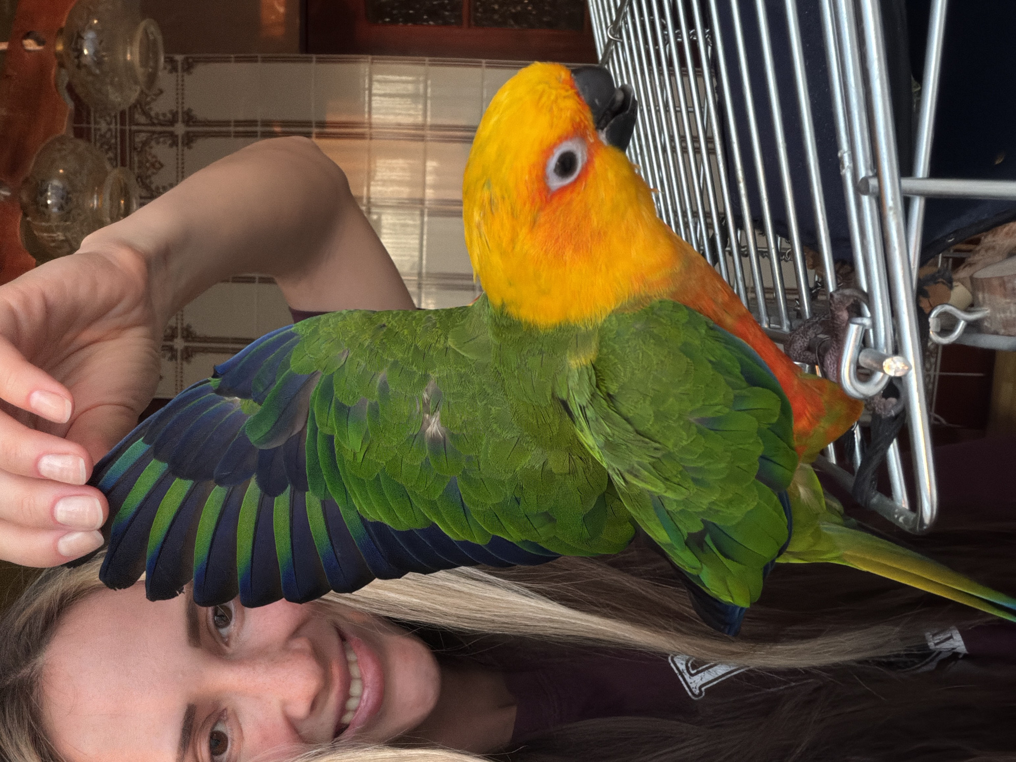 A woman with blonde hair is holding a vibrant, multi-colored parrot in her hands. The parrot has a yellow head, a green body, and blue-tipped wing feathers. The woman is smiling at the camera. A birdcage is partially visible in the background. The location is indoors, likely in a home. There is no indication of the location being in Khimki, Russia.