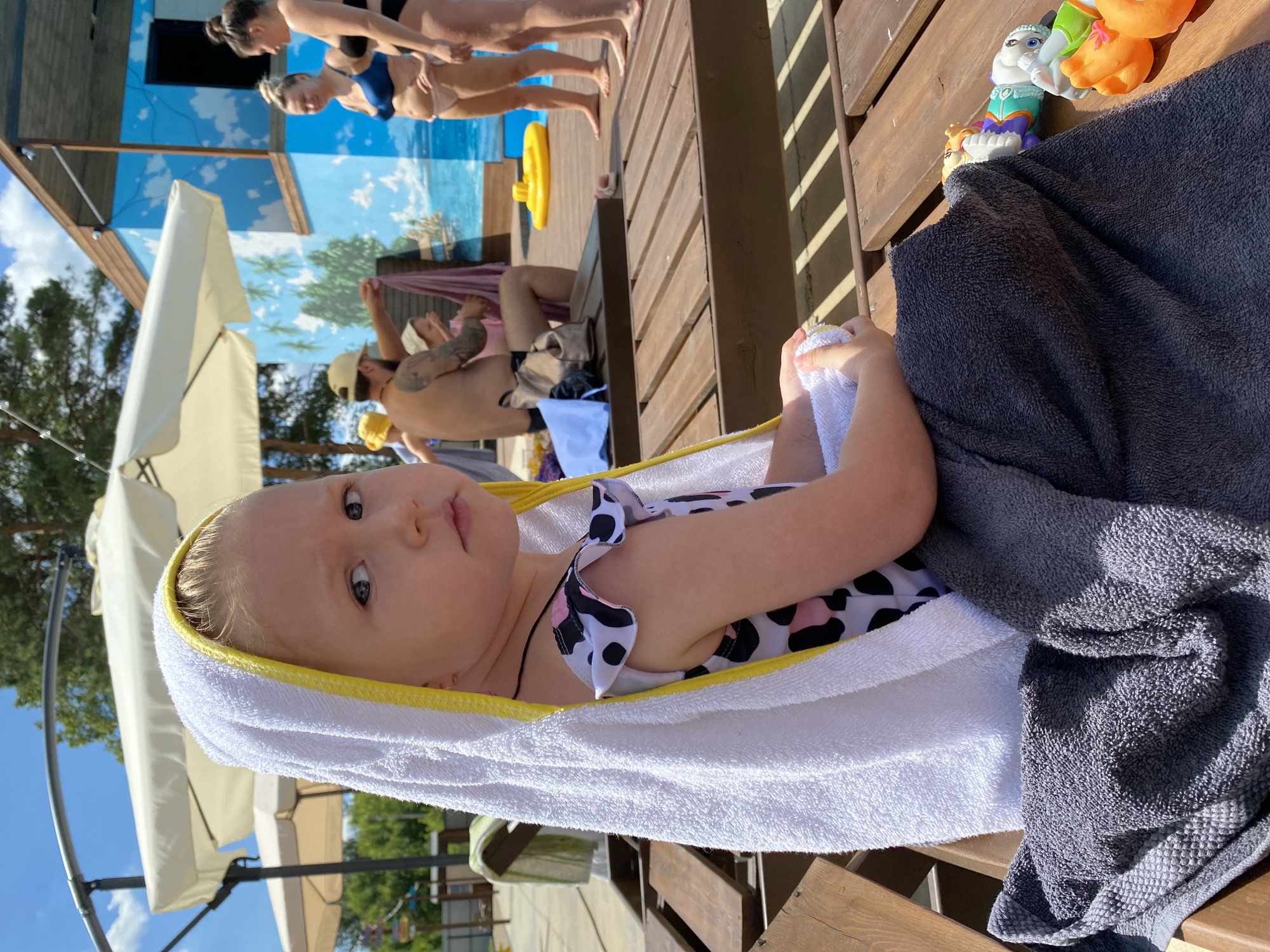The image shows a baby wrapped in a white towel with a yellow trim, lying on a wooden deck. The baby is wearing a black and white spotted swimsuit and looking at the camera with a curious expression. Next to the baby, a dark gray towel is draped over the wooden surface. In the background, two women in swimwear are visible near a building with a mural of a blue sky and palm trees. Another person, likely a man wearing a cap, is partially visible in the middle ground. Several beach umbrellas and trees are also present, suggesting an outdoor setting. Toys, including a white and orange plush toy and a small yellow inflatable ring, are scattered on the deck. The scene appears to be set during the daytime, with bright sunlight casting shadows on the wooden planks, indicating fair weather. The overall atmosphere suggests a relaxed day at a pool or beach resort. No visible text is present in the image.