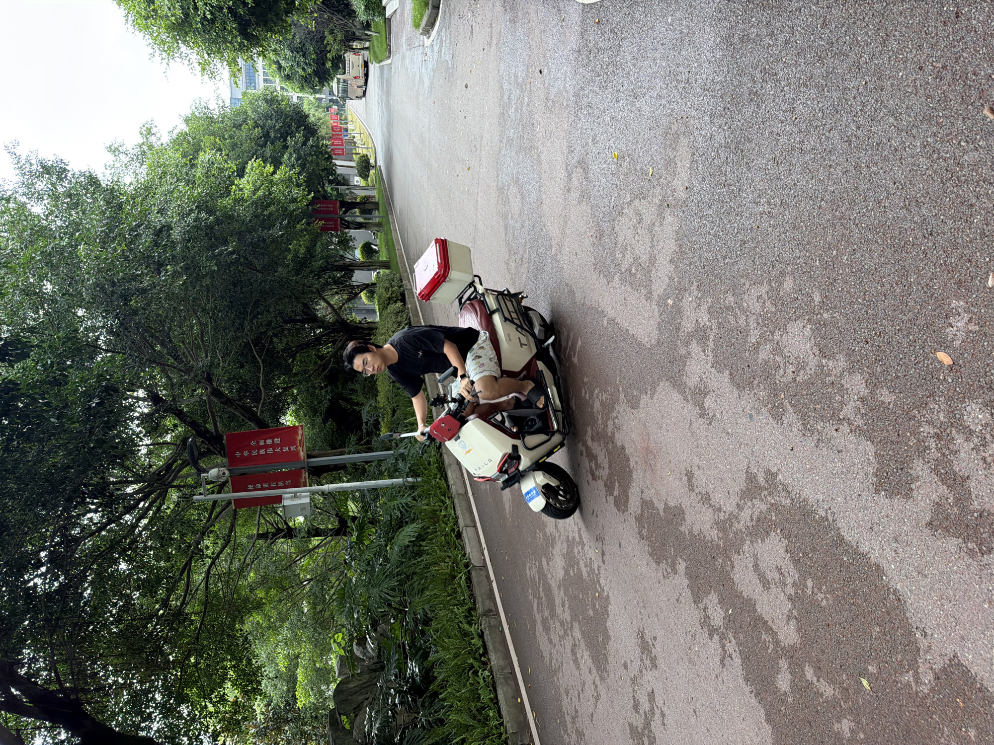 A young East Asian male wearing glasses, a black t-shirt, and patterned shorts rides a white and red electric scooter on a wet asphalt road. He is positioned in the left half of the frame, facing forward, with his hands on the handlebars. The scooter has a red and white storage box mounted on the rear. The road is lined with trees on both sides, and red banners are visible hanging from poles along the left side of the road. In the distance, a white bus is visible. The environment appears to be a park or campus setting, possibly in the city of Jiangjin, China, given the context provided. The overall lighting suggests an overcast day.