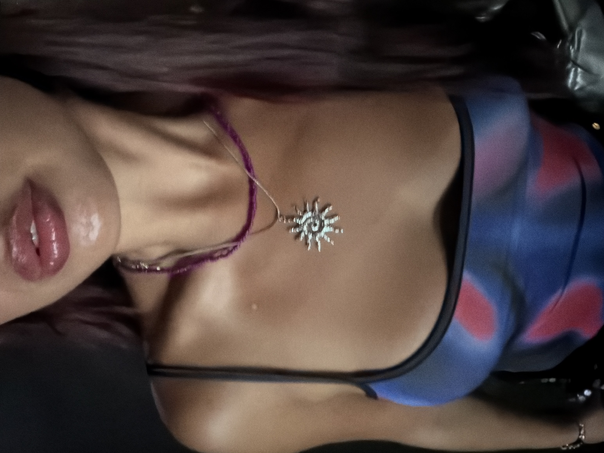 The image shows a close-up of a woman's chest and lips. She has on a blue and pink tie-dye tank top and two necklaces, one with a sun pendant. Her lips are glossy. The background is dark and out of focus. There is no indication of the location being in Arugam Bay, Sri Lanka.